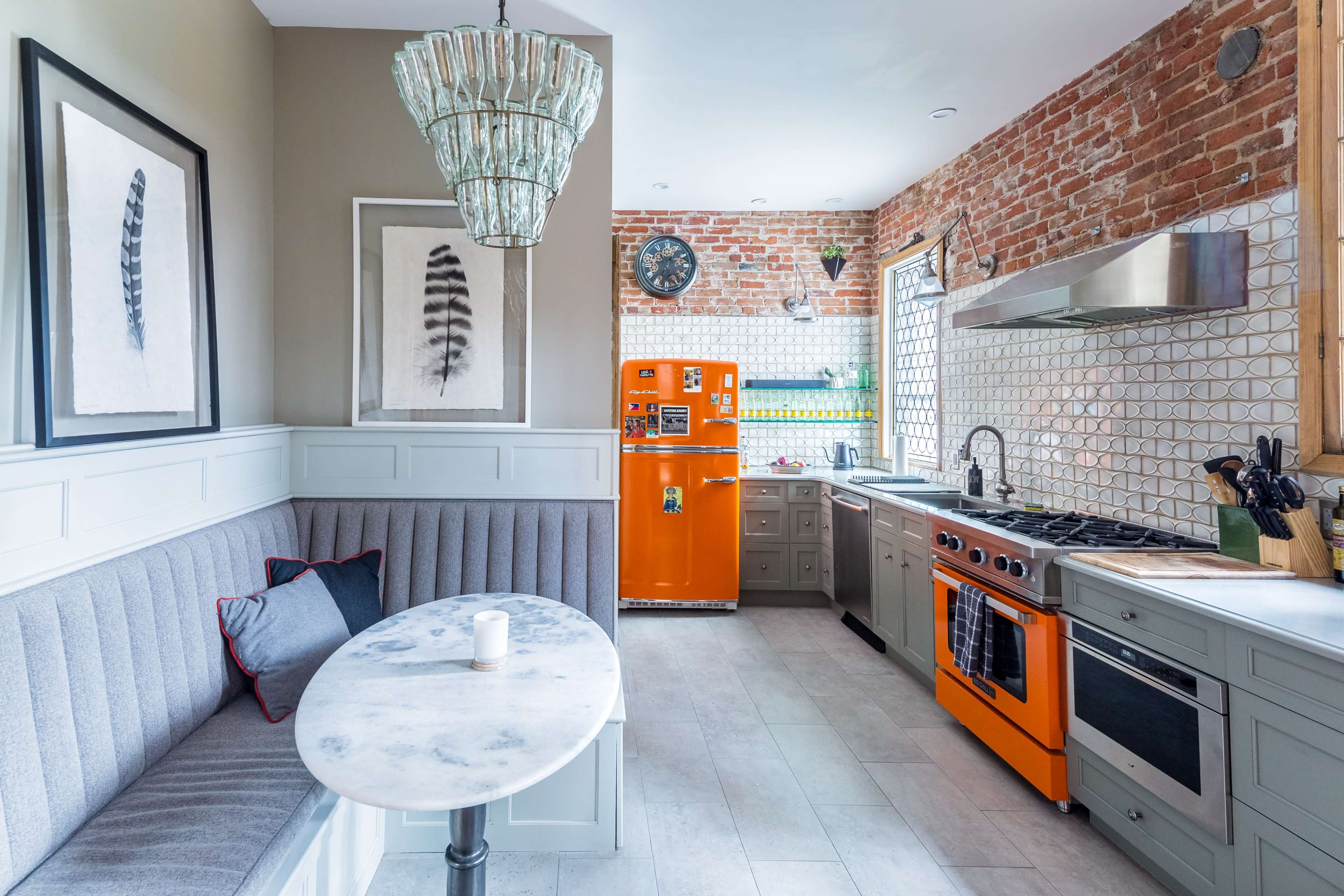 The kitchen features a brick wall, an orange refrigerator, and a marble-topped table with a gray upholstered seating area.
