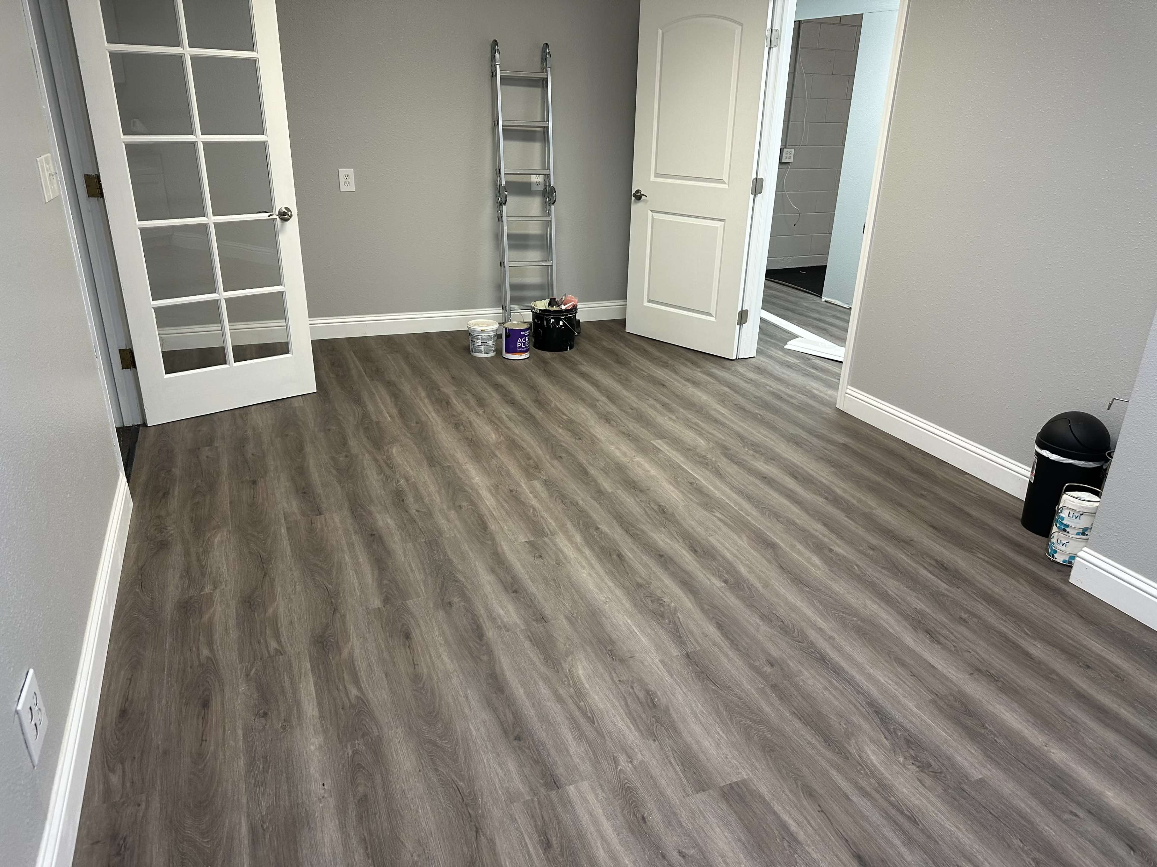 A vacant room with laminate flooring, two doors, a ladder in the corner, and paint cans placed on the floor.