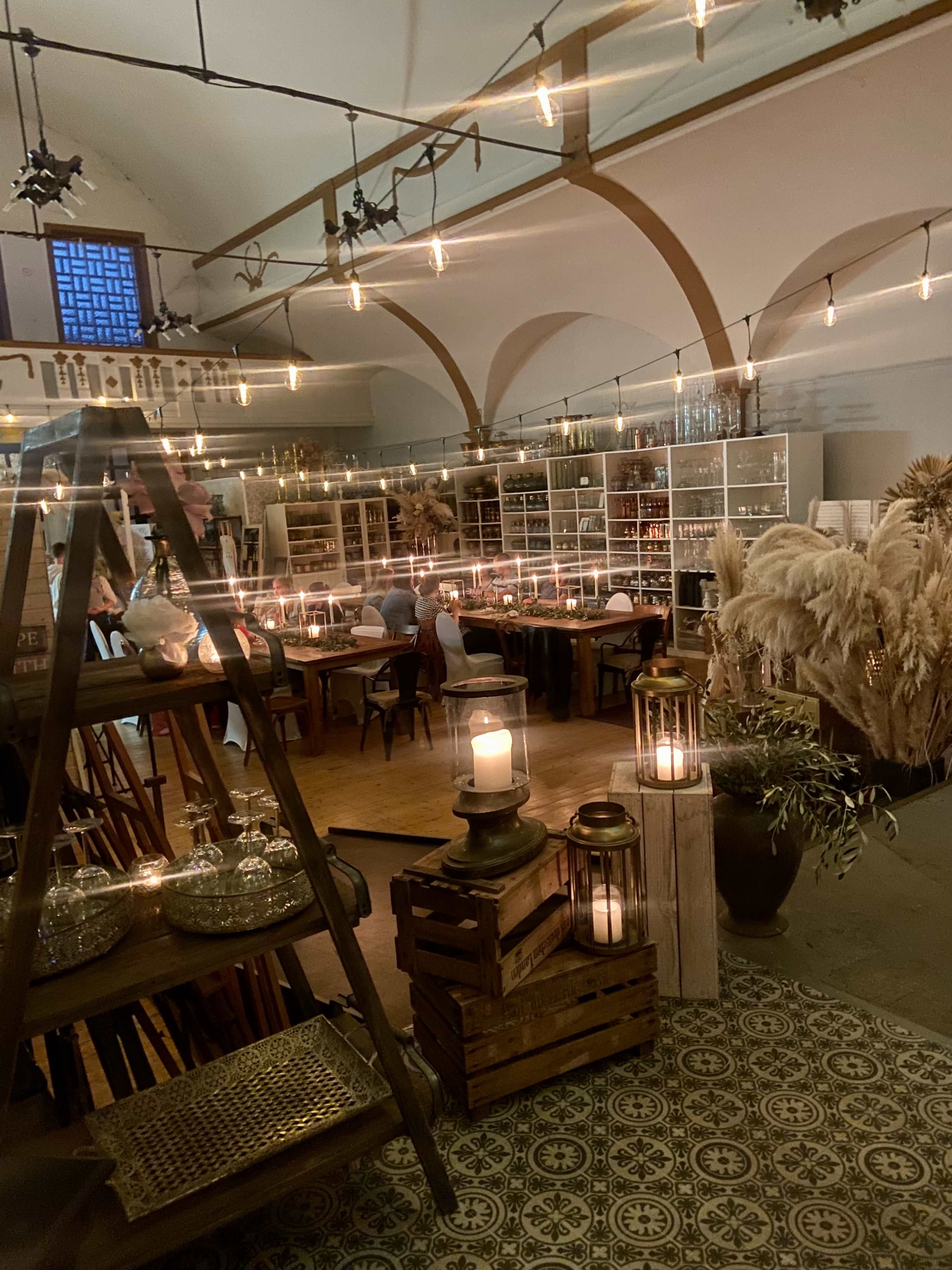 The image shows a warmly lit interior space featuring wooden tables, decorative plants, and a variety of glassware arranged on shelves.