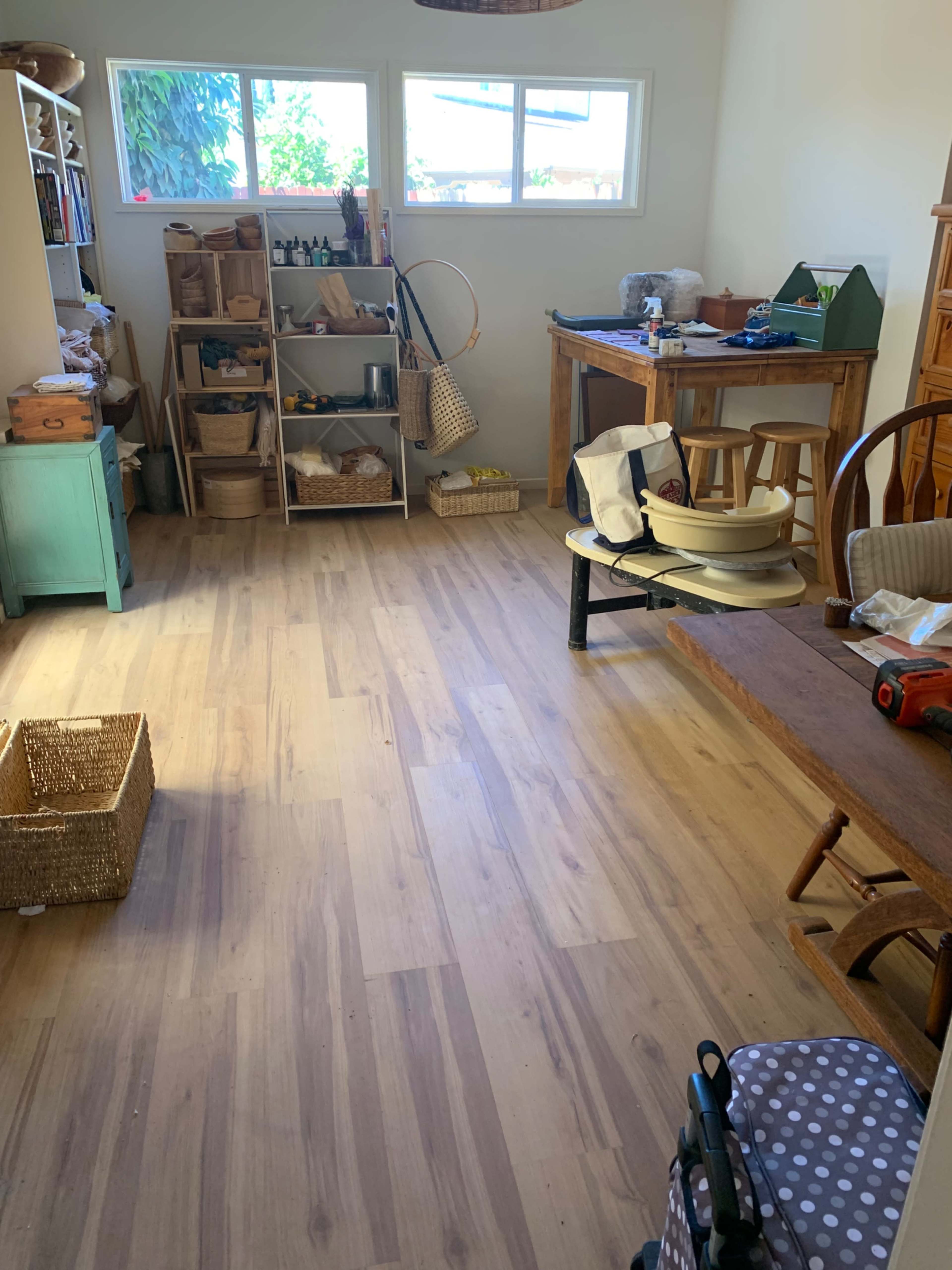 The image shows a spacious, light-filled room with wooden flooring, shelves filled with various items, and furniture including a table and chairs.