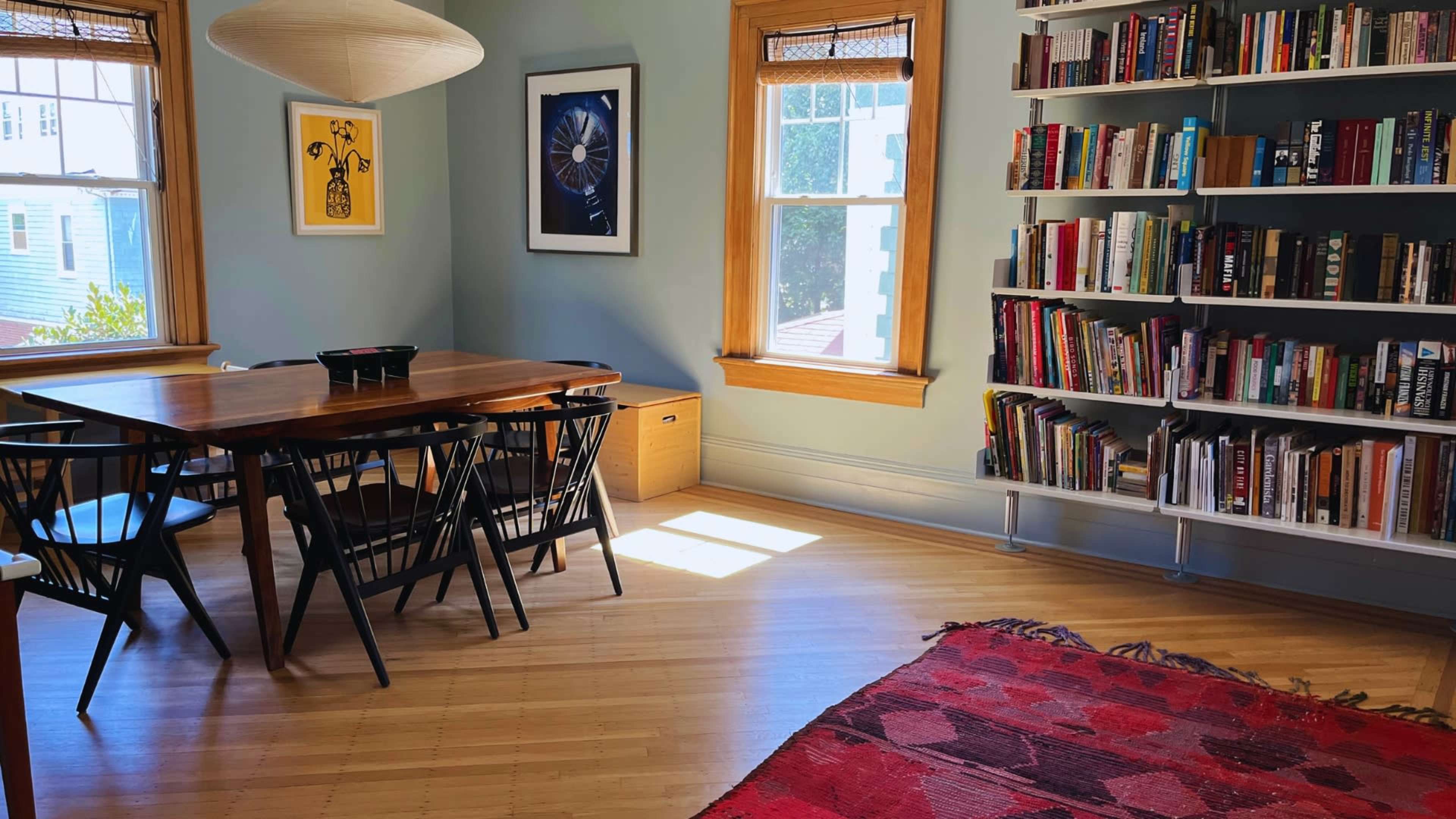 The image depicts a well-lit room featuring a wooden dining table surrounded by chairs, a bookshelf filled with books, and a patterned rug on the floor.