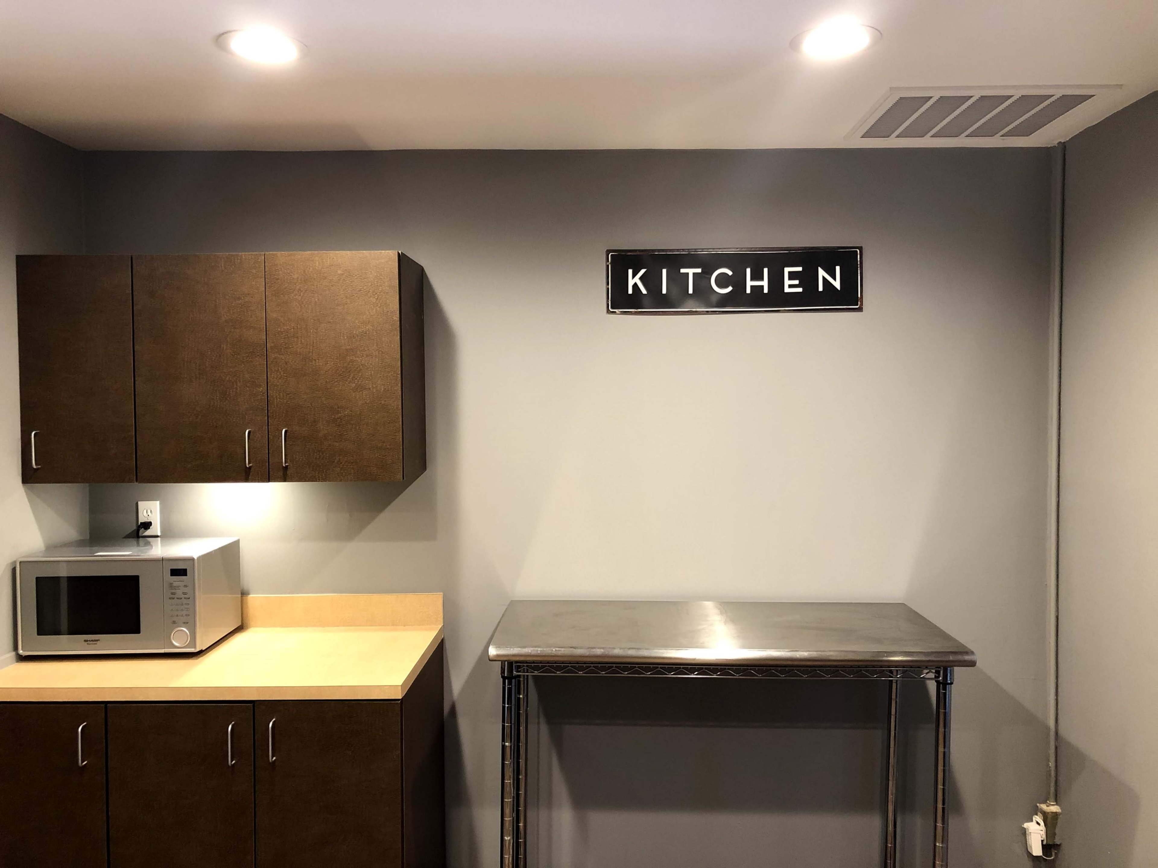 The image shows a kitchen area featuring brown cabinets, a microwave, and a metal countertop below a sign that reads "KITCHEN."
