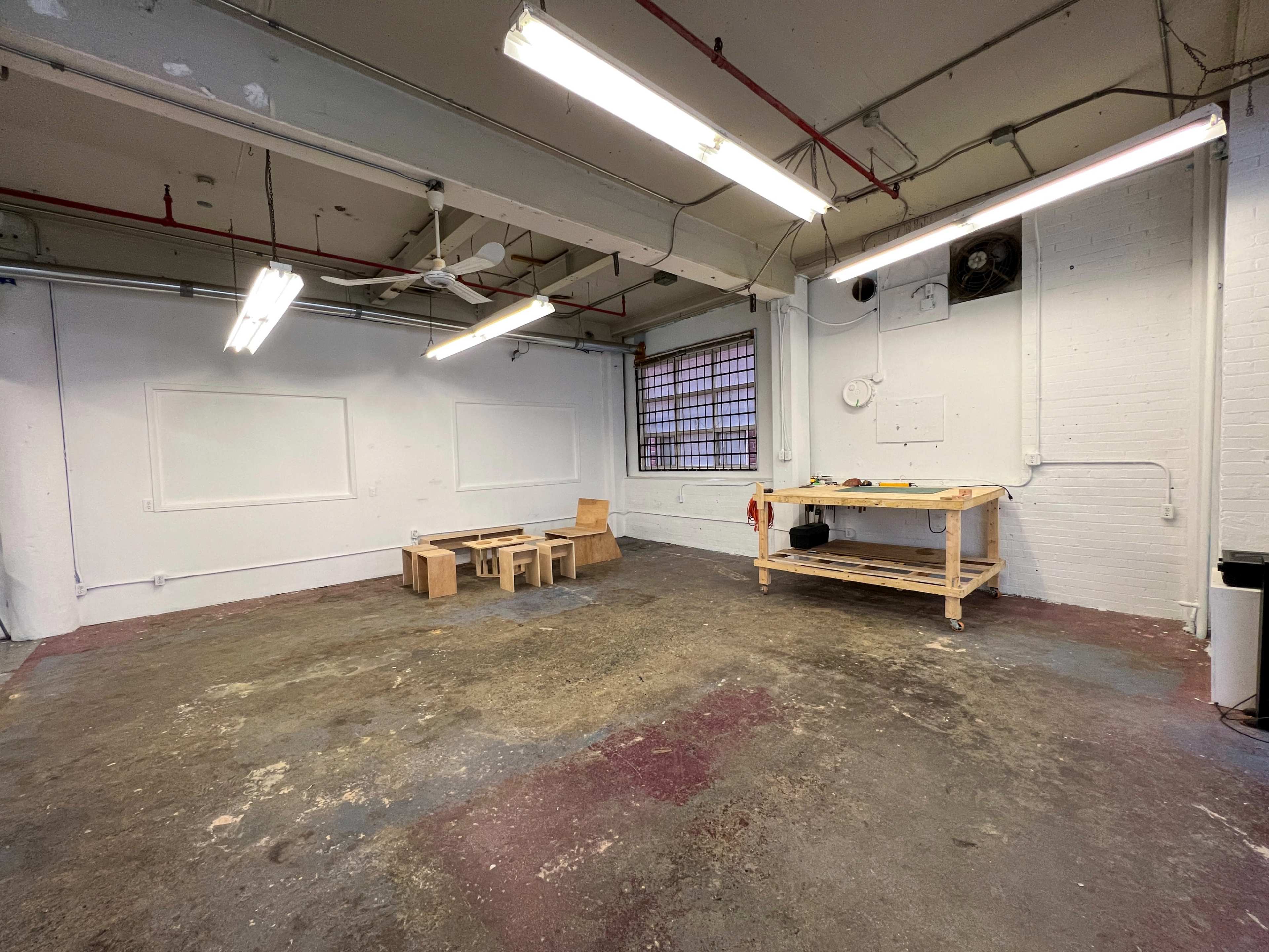 An empty industrial workspace with a wooden table, a few chairs, and visible ductwork along the ceiling.