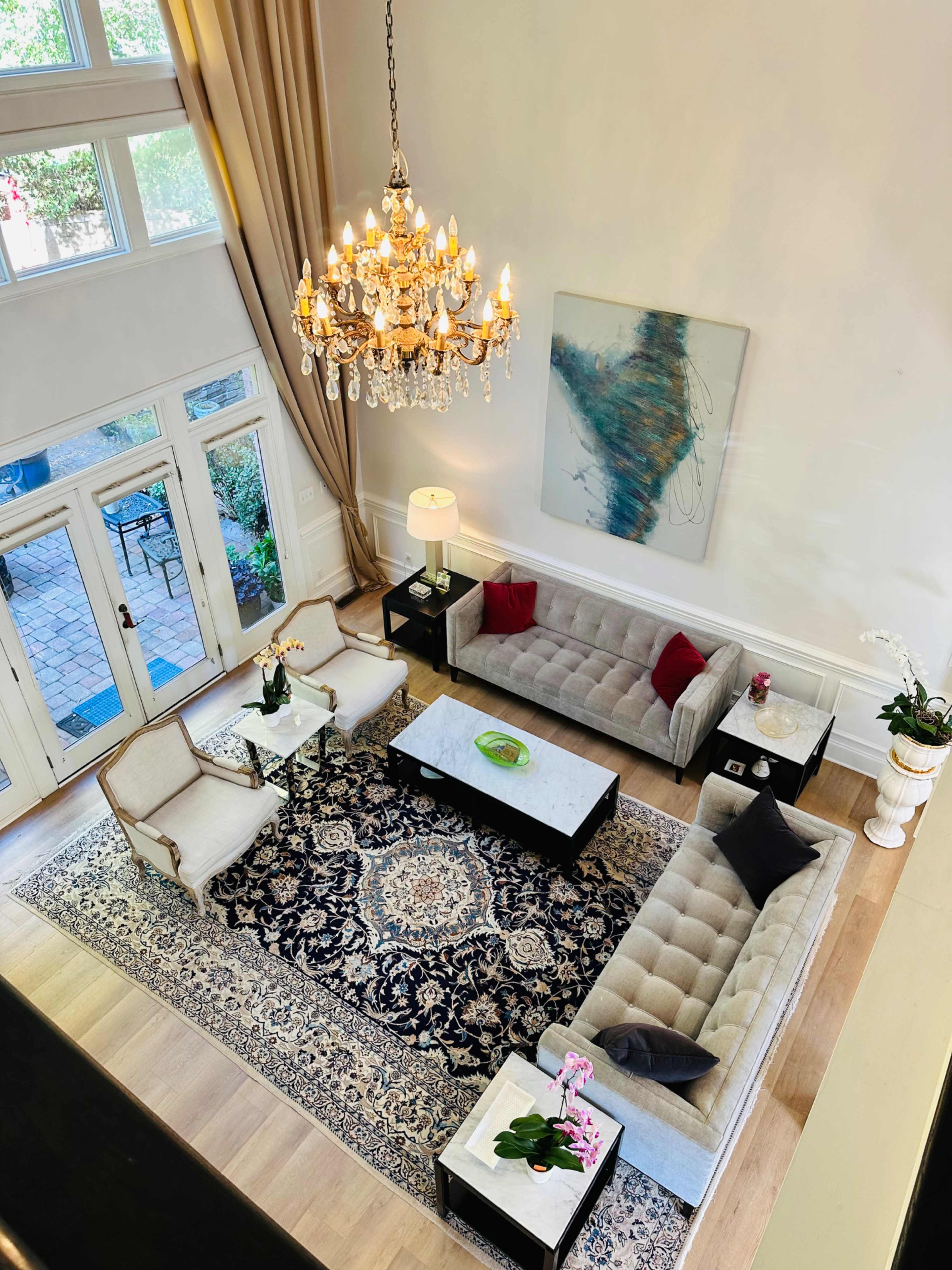 The image shows a stylish living room with a chandelier, two sofas, a coffee table, and a large area rug, characterized by high ceilings and ample natural light.