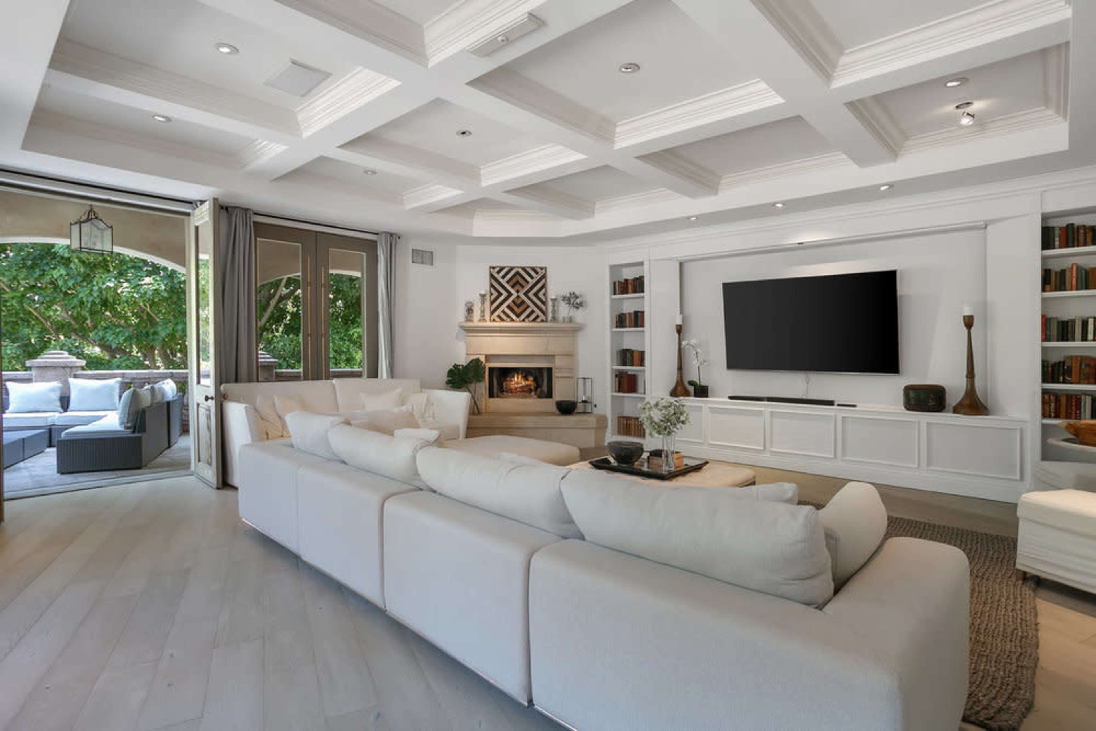 The image shows a spacious living room with a white sectional sofa, a fireplace, a television mounted on a wall, and large windows overlooking a patio.