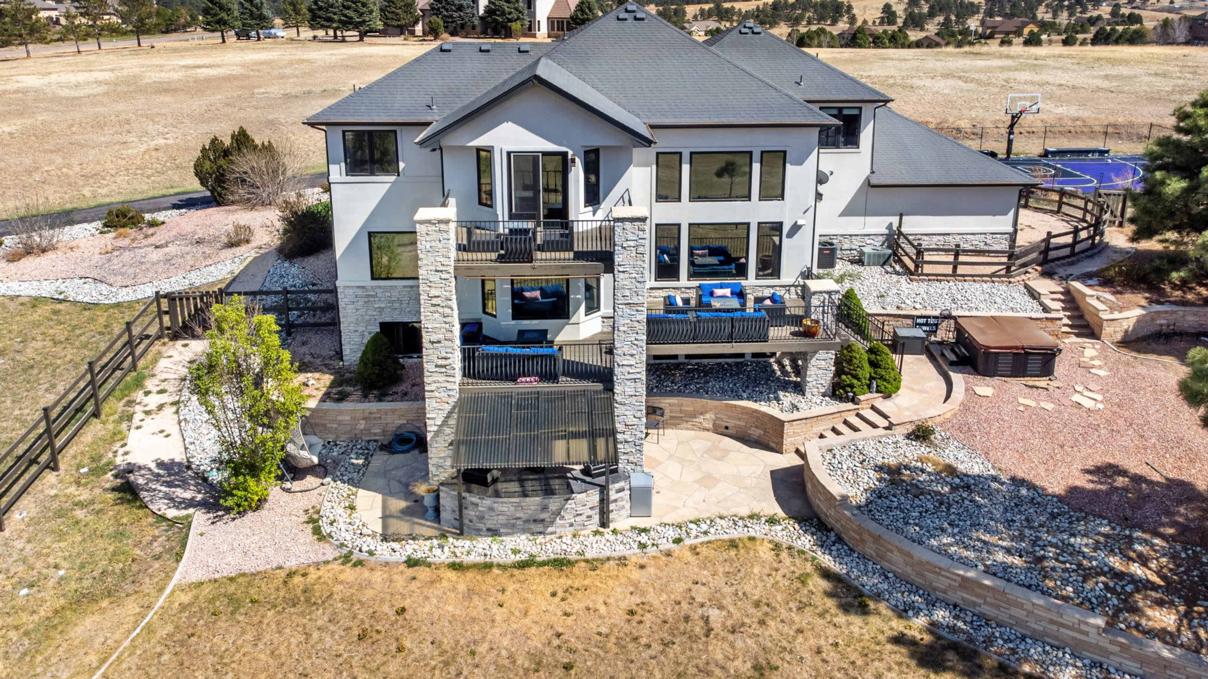 The image shows a large, modern two-story house with a stone-covered patio and a hot tub, set against a backdrop of an open landscape.