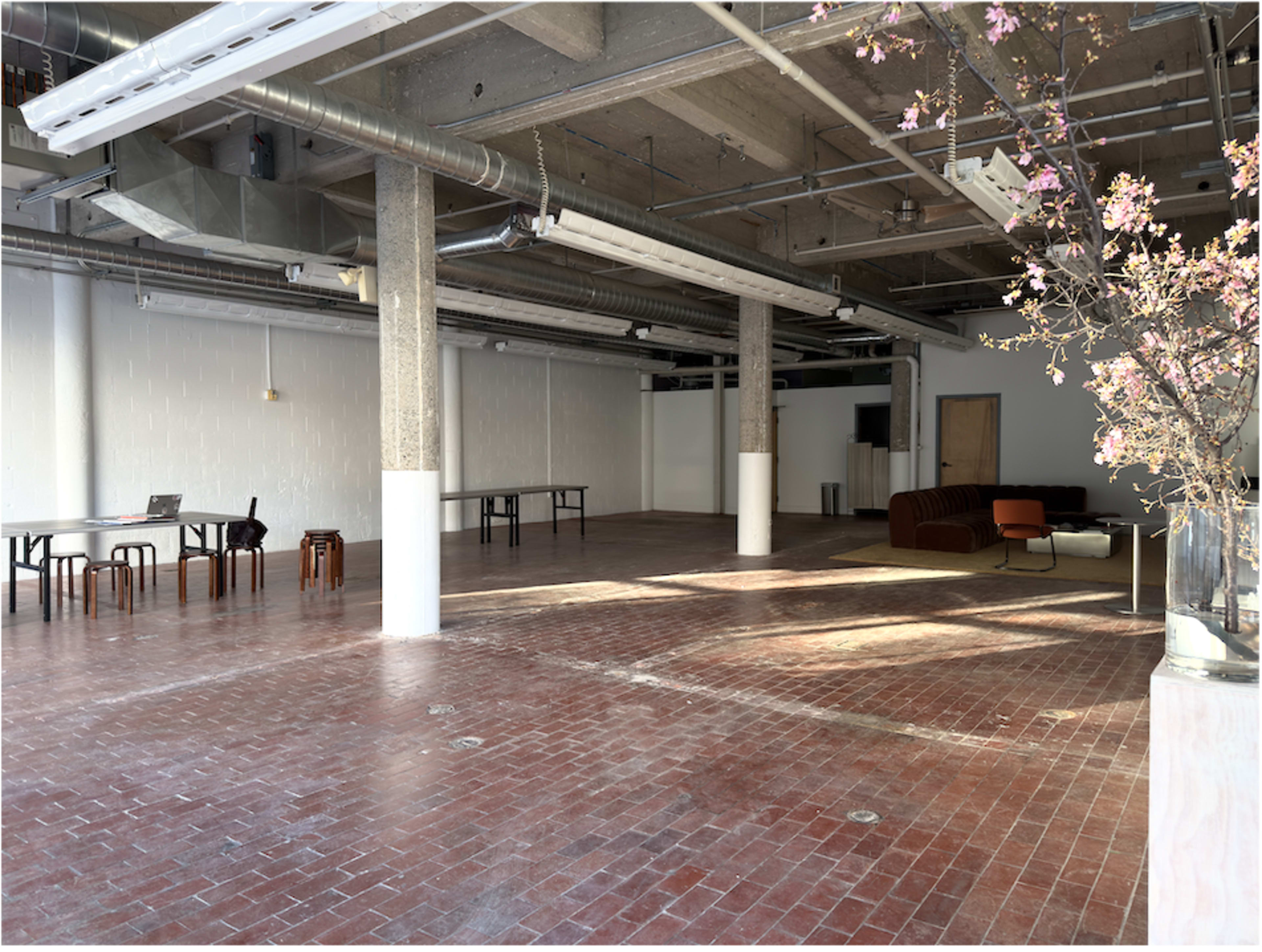 The image shows a spacious, industrial-style interior with exposed concrete ceilings, brick flooring, and minimal furniture, including a couch and tables.