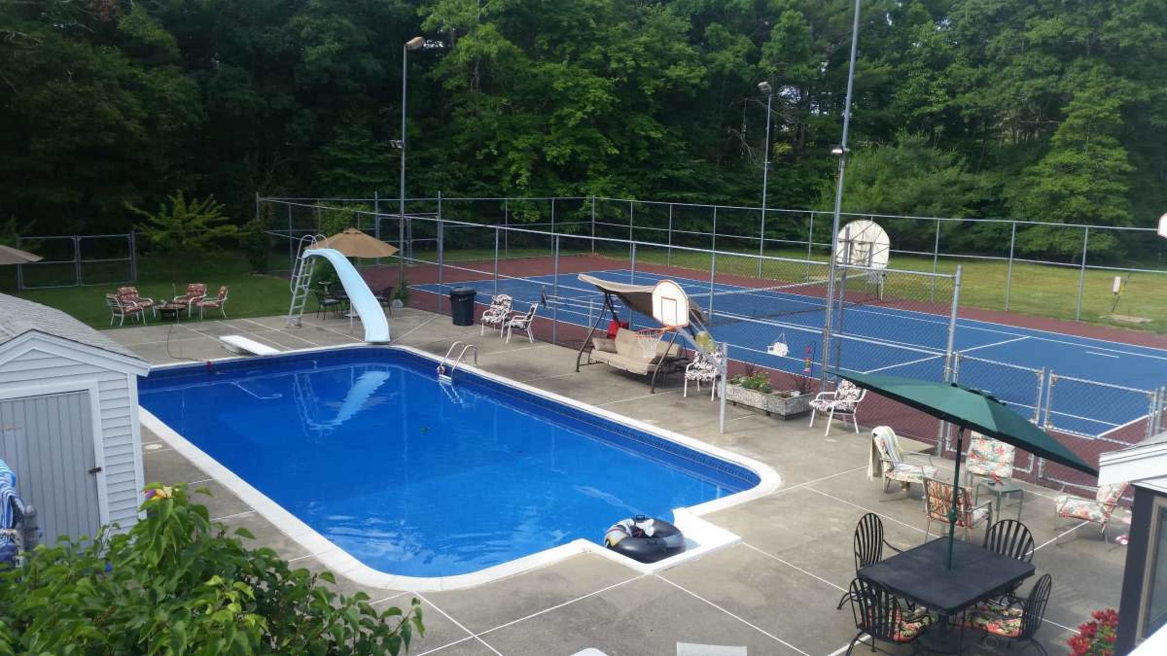 The scene features a swimming pool with a slide surrounded by lounge chairs, next to a tennis court fenced in by trees.