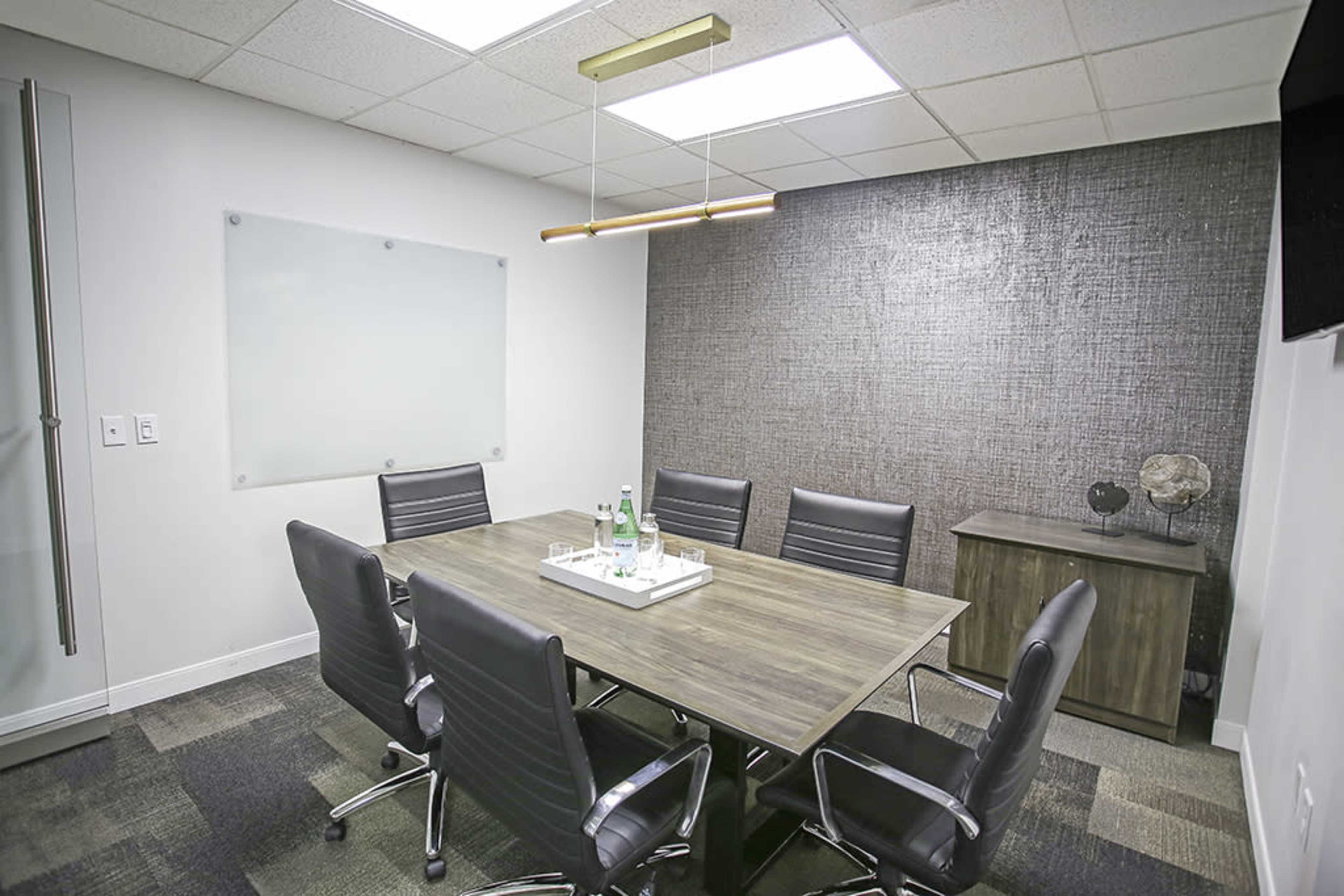 A modern conference room features a rectangular wooden table surrounded by black leather chairs, with a glass whiteboard on one wall and a textured accent wall.