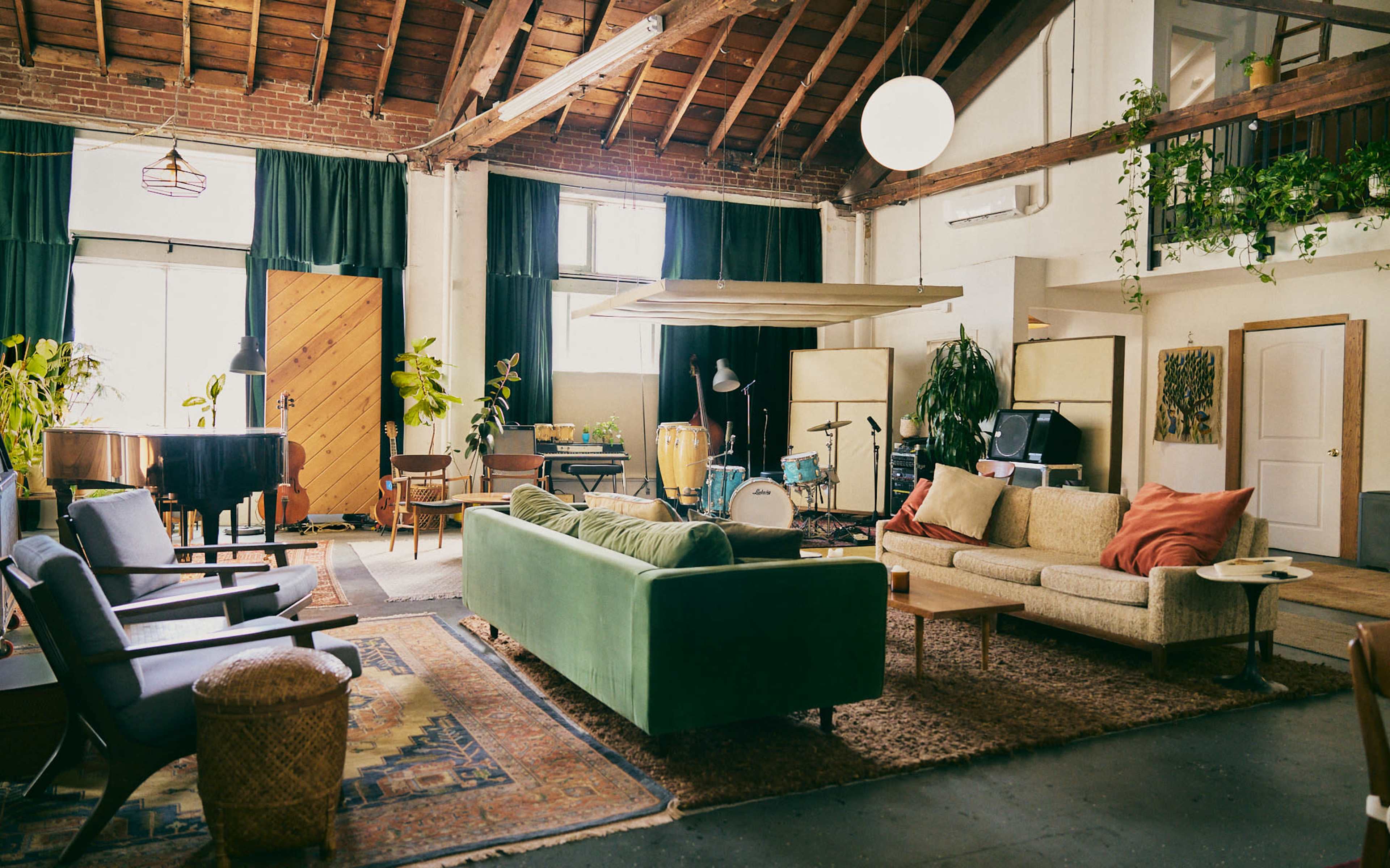 The image features a spacious, well-lit loft living area with exposed wooden beams, a green sofa, various seating arrangements, a piano, and musical instruments in the background.