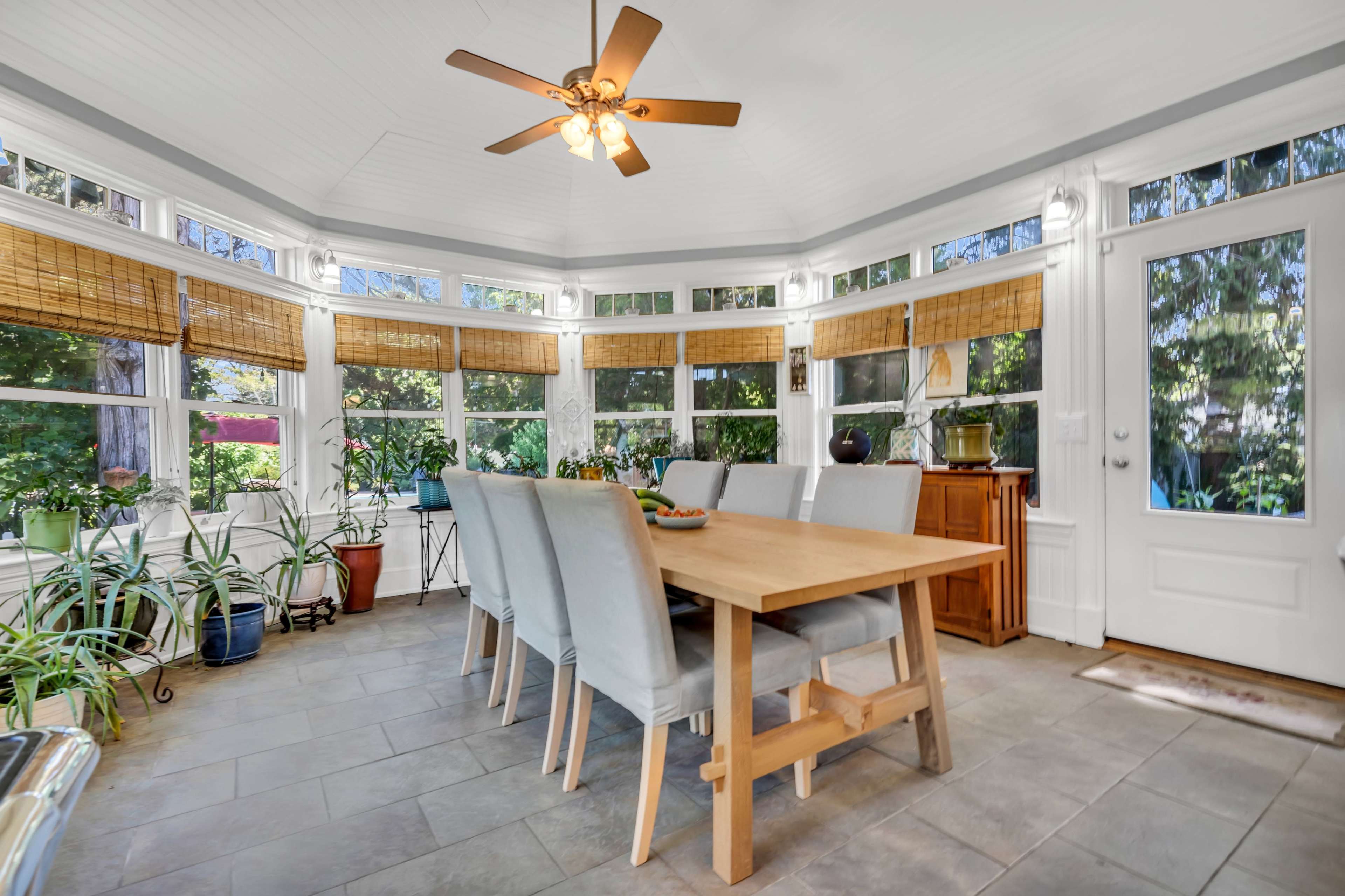 The image shows a sunlit dining area with a wooden table surrounded by upholstered chairs, set in a room featuring large windows and bamboo shades.
