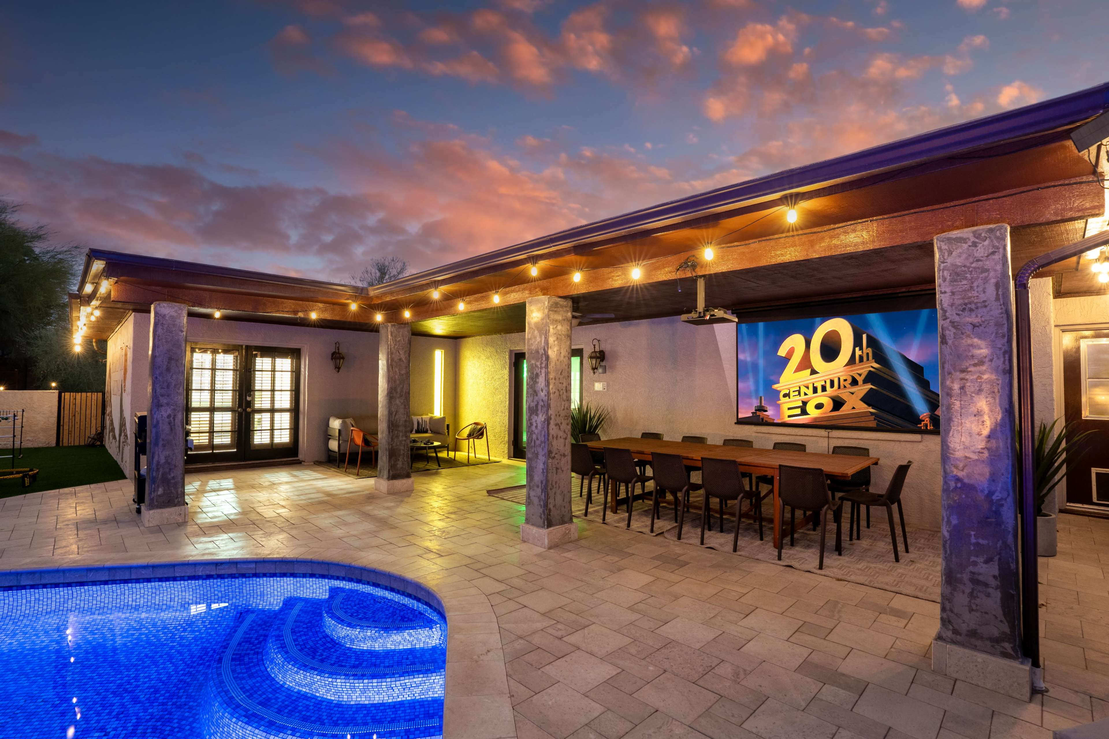 A modern outdoor entertainment area features a pool, a dining space, and a large screen displaying a movie logo against a twilight sky.