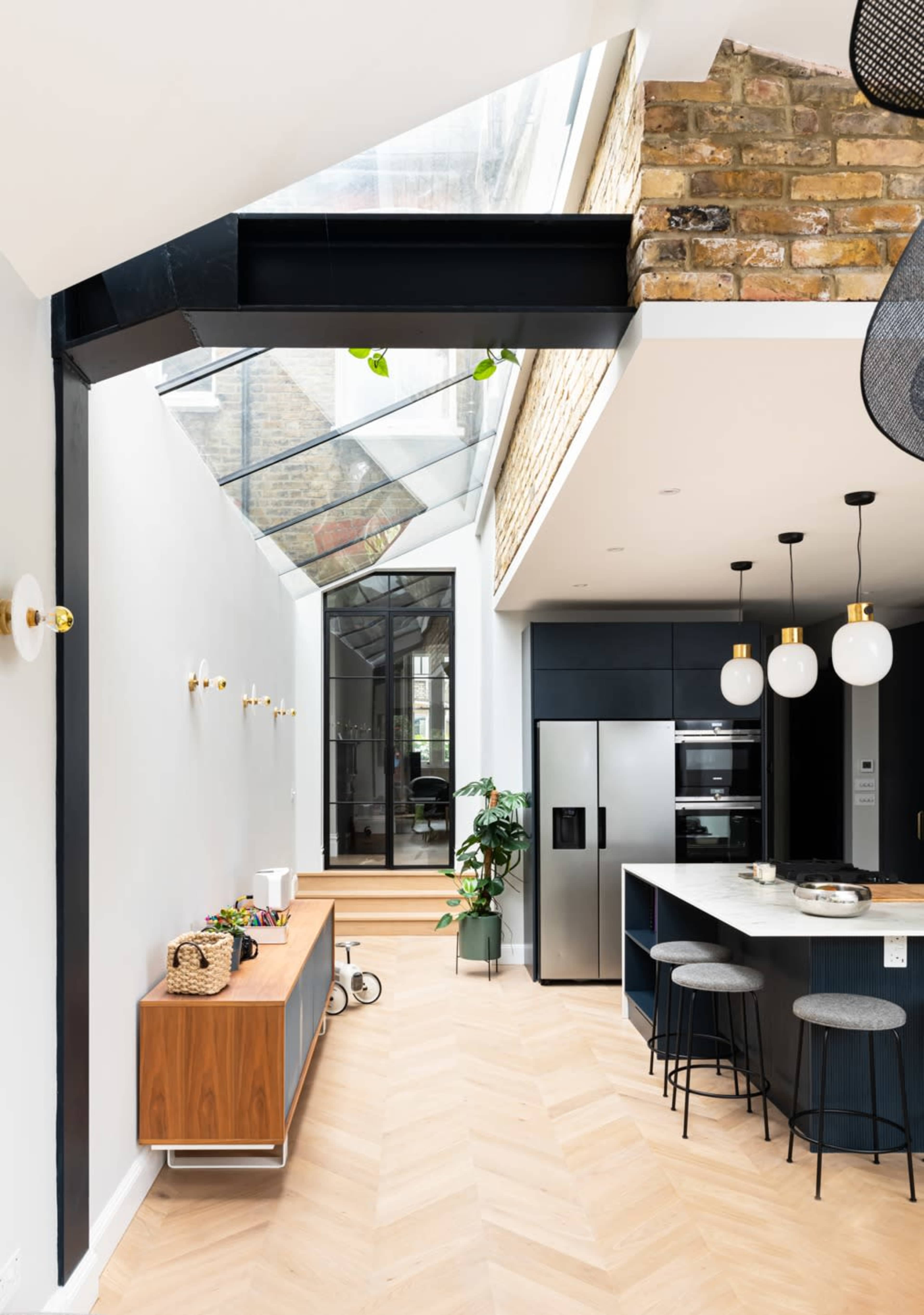The image shows a modern kitchen with a glass roof, a central island, and a combination of dark cabinetry and light wood flooring.
