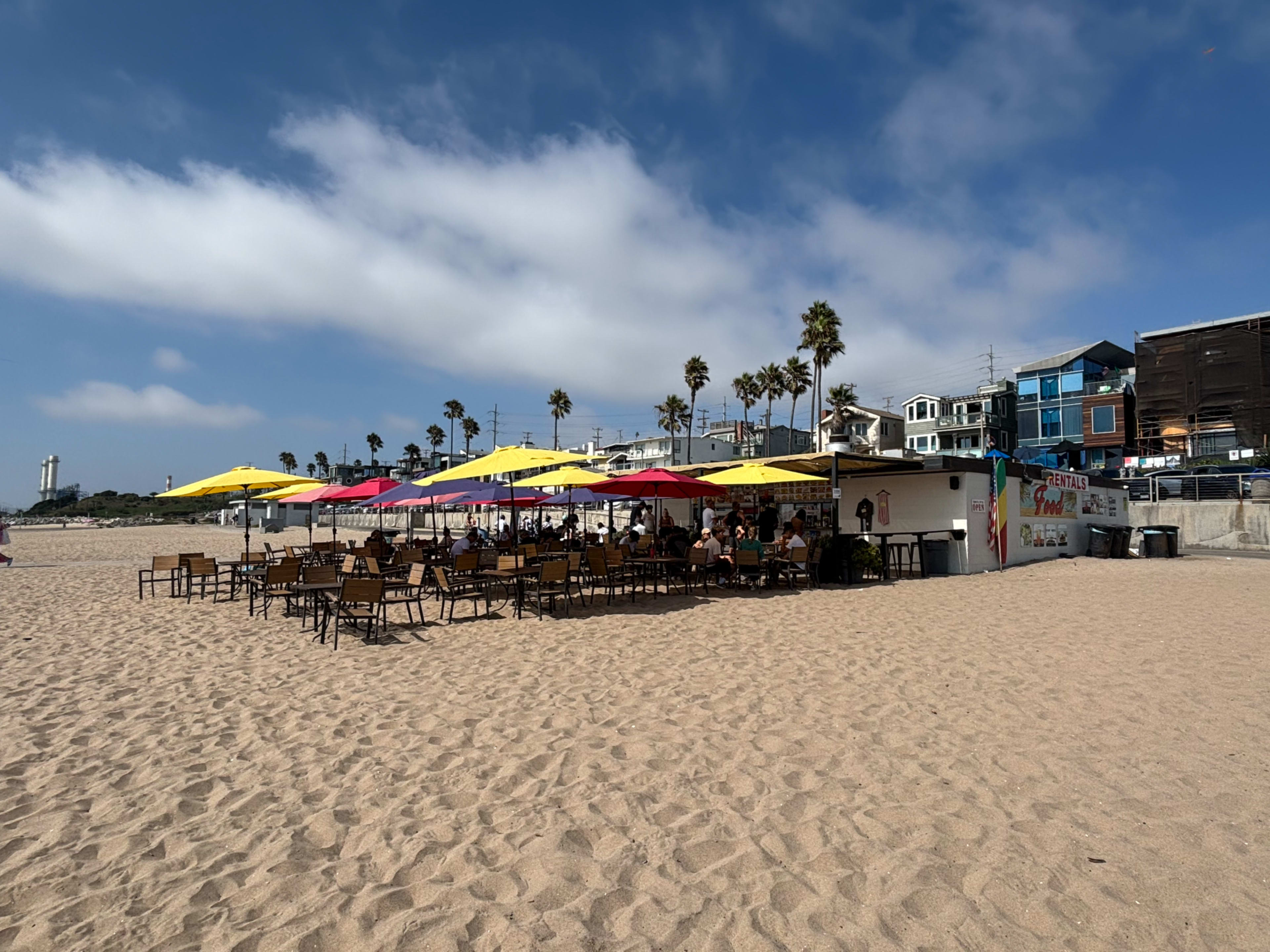 A beachside café with colorful umbrellas provides outdoor seating on sandy shores, surrounded by palm trees and coastal buildings.