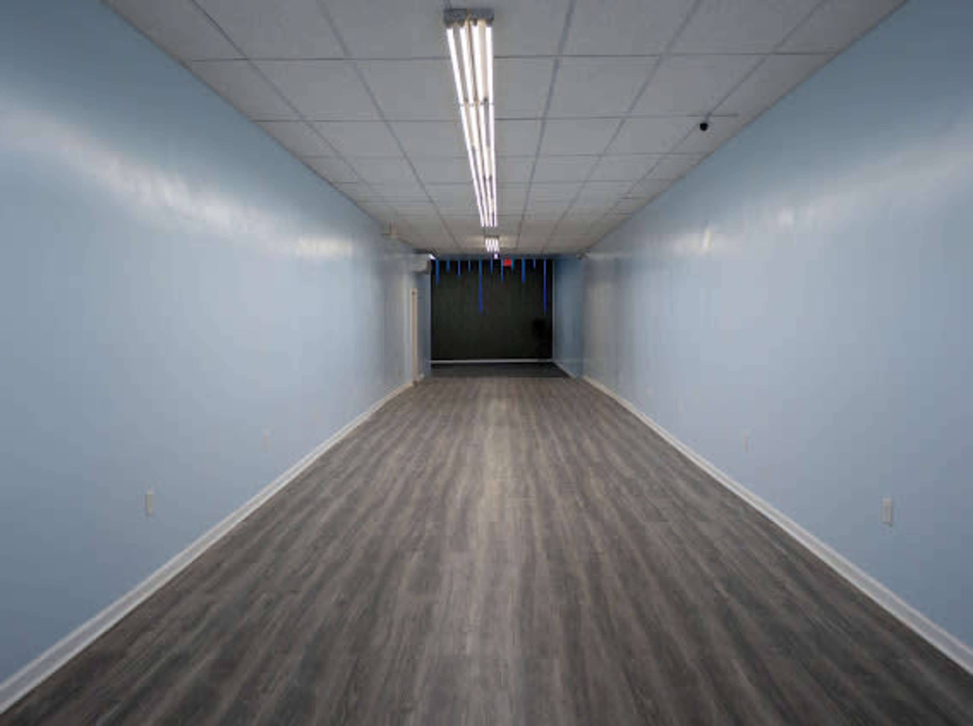 The image shows a long and empty hallway with a light blue wall and gray wood-like flooring.