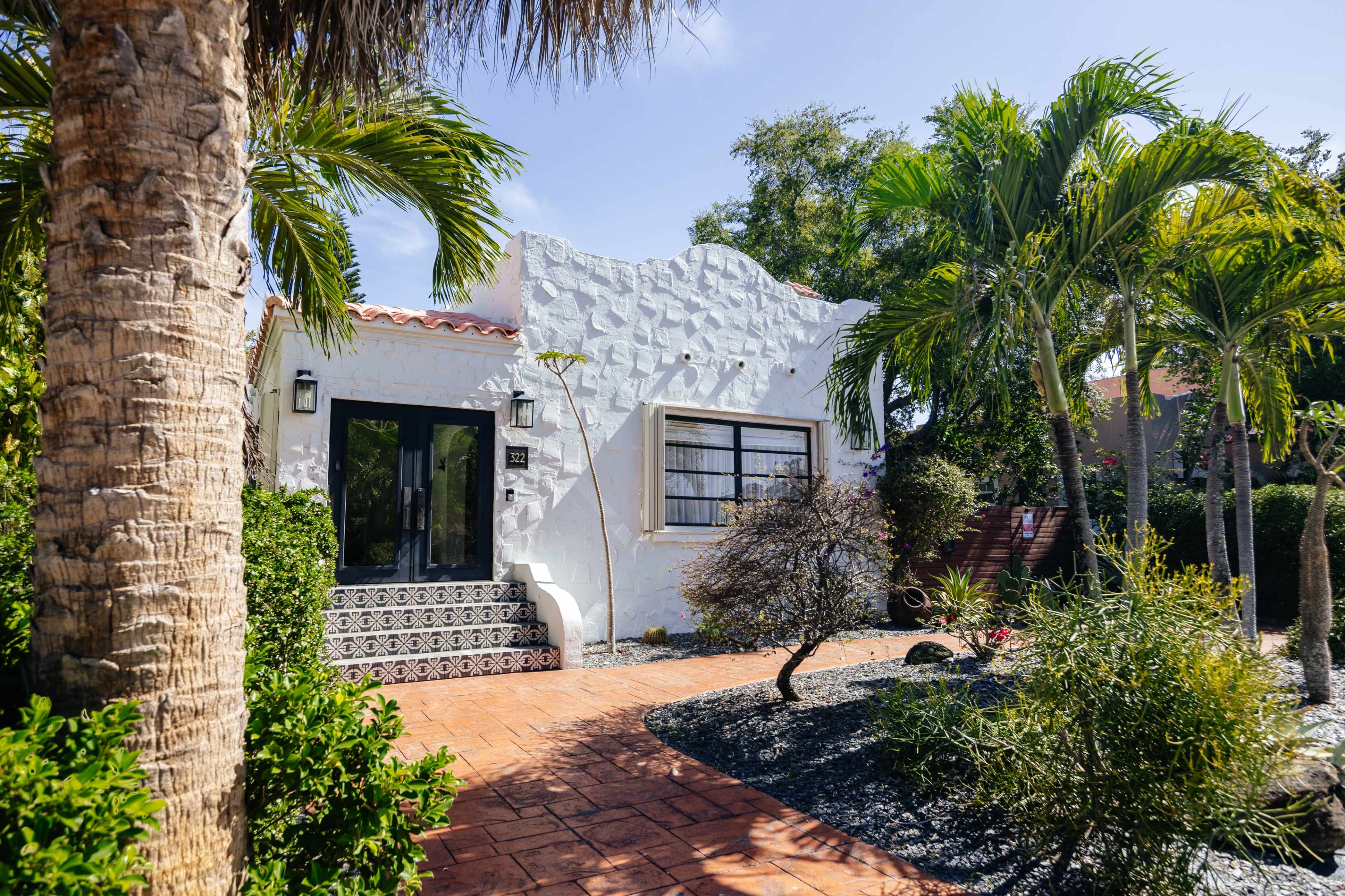 A white stucco house with a patterned entryway is surrounded by tropical plants and palm trees.