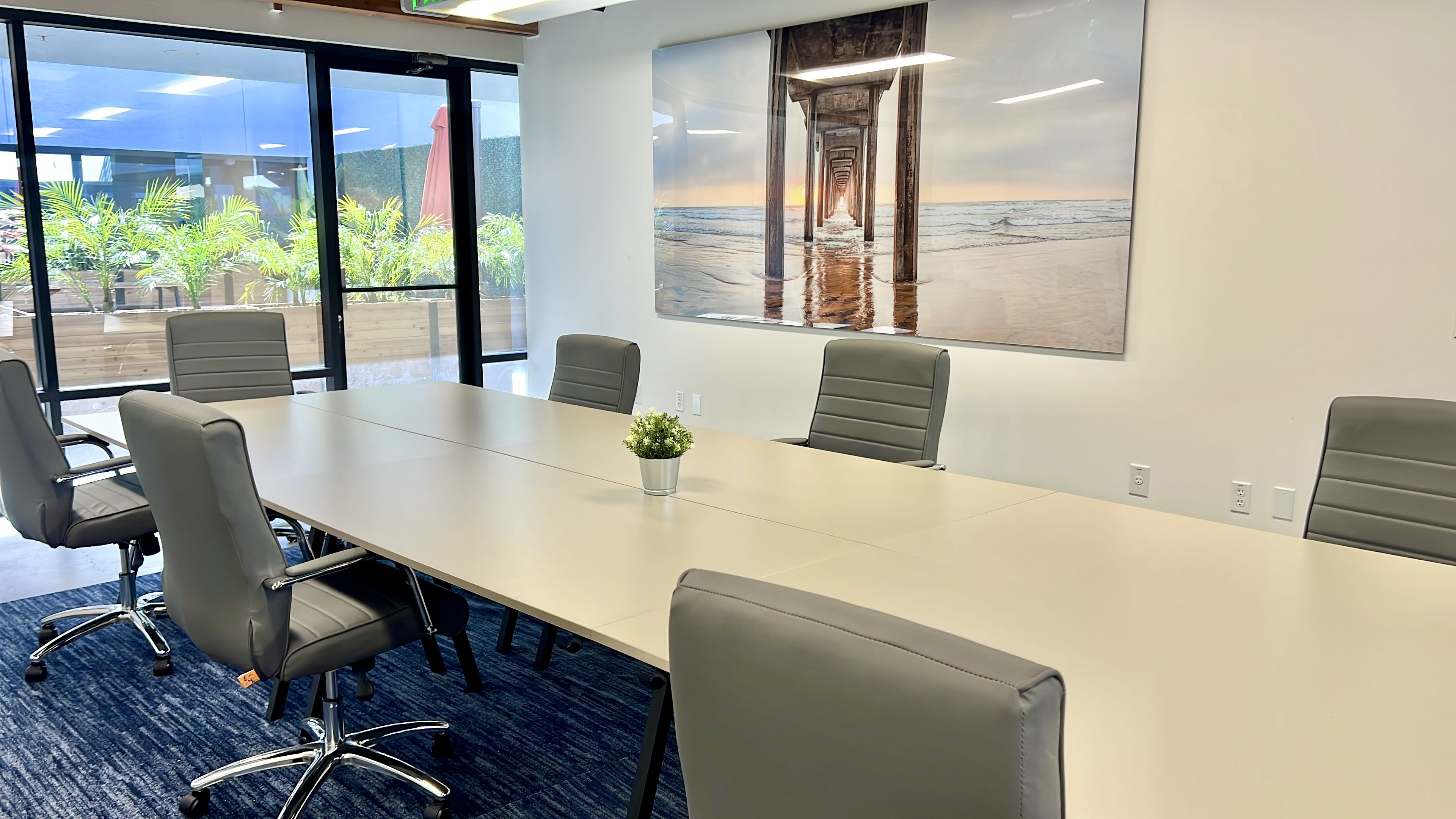 A modern conference room features a large rectangular table surrounded by ergonomic chairs, with a beach-themed artwork on the wall and large glass doors leading to an outdoor area.