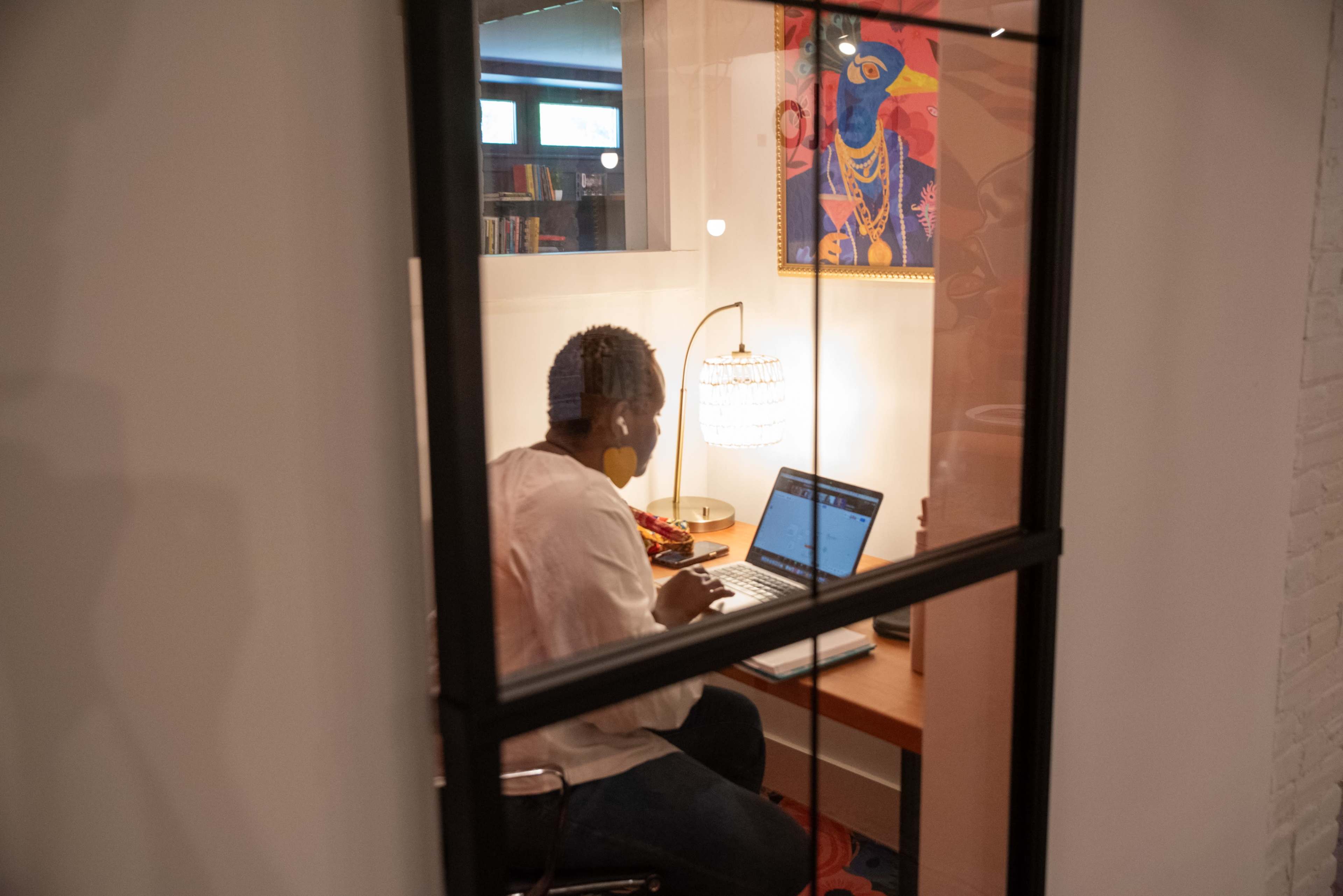A person is seated at a desk working on a laptop inside a well-lit office space, featuring a decorative lamp and colorful artwork on the wall.