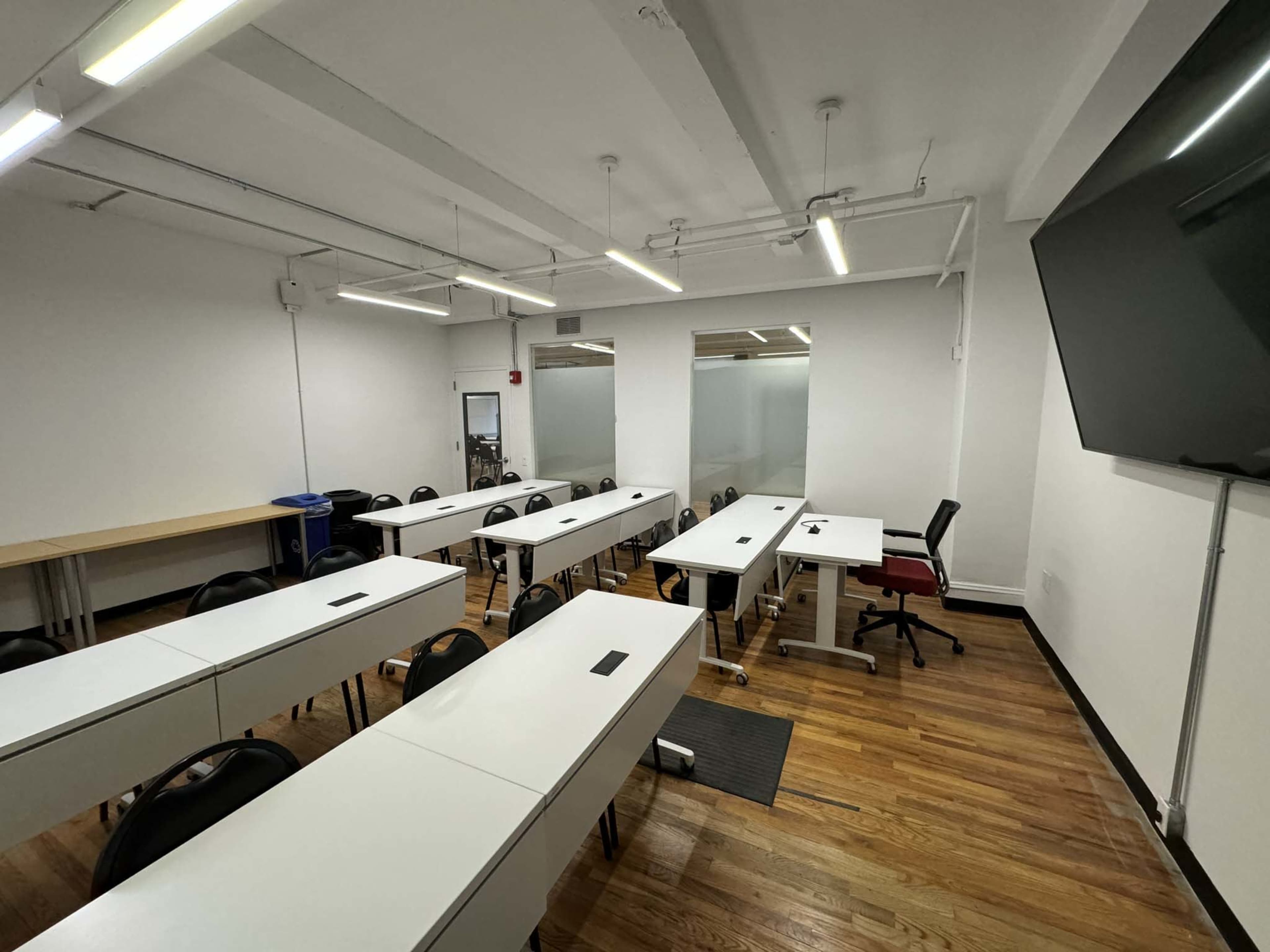 A spacious classroom features several rows of desks and chairs, with a large screen and a wooden table along one wall.