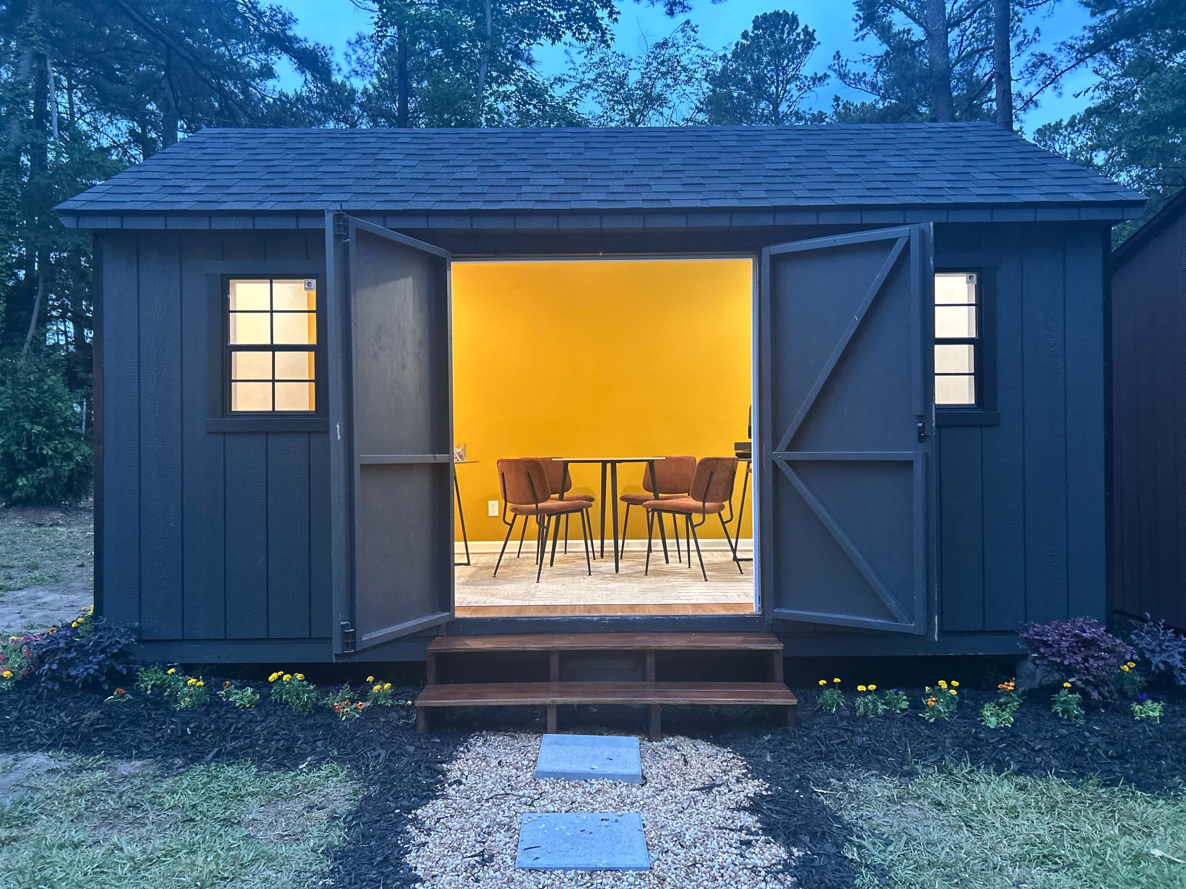 A small gray shed with open doors reveals a brightly lit interior featuring a yellow wall, two chairs, and a small table.