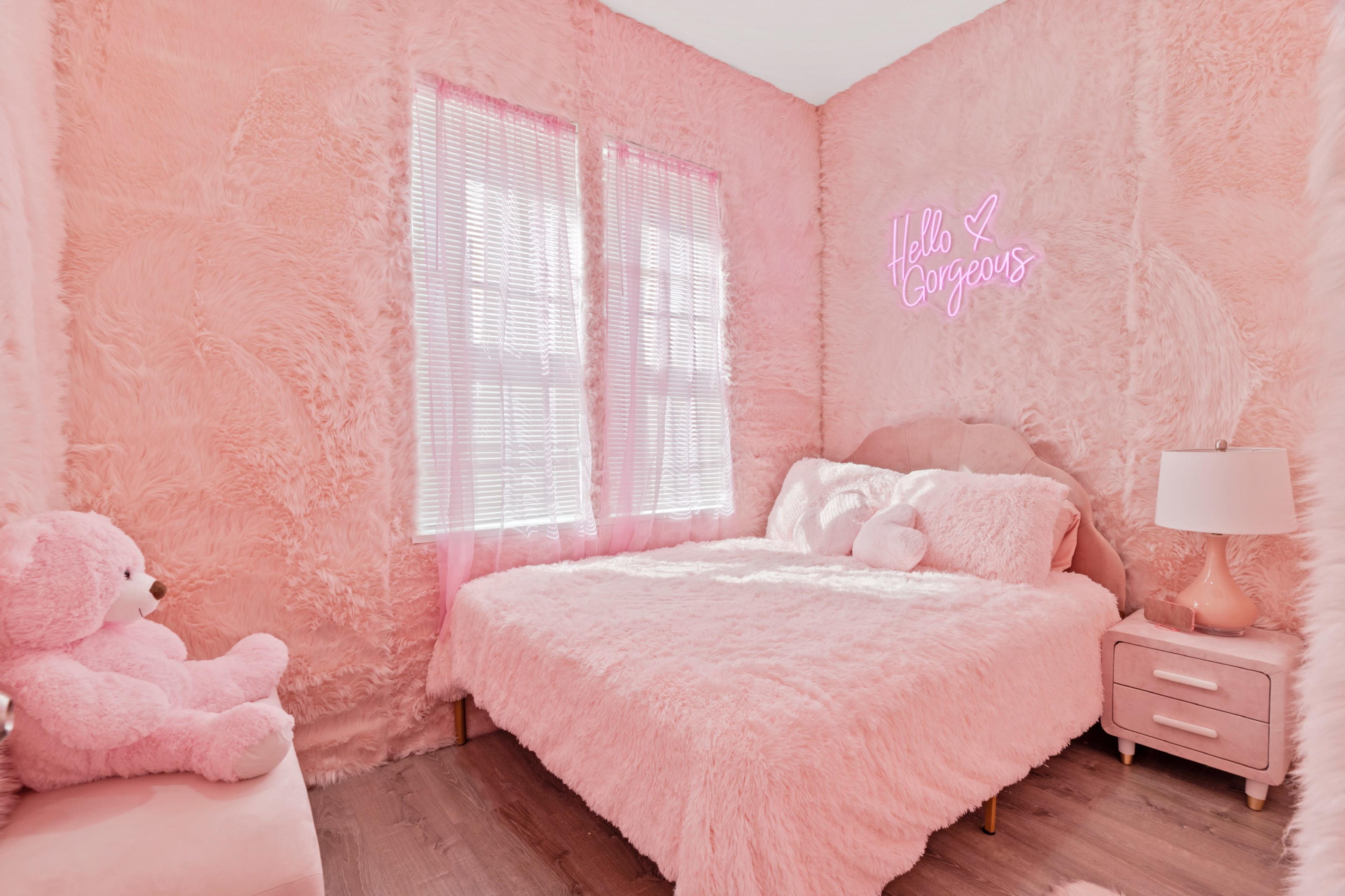 A pink, plush-furnished bedroom features a bed with fluffy blankets, a neon sign, and a teddy bear in the corner.