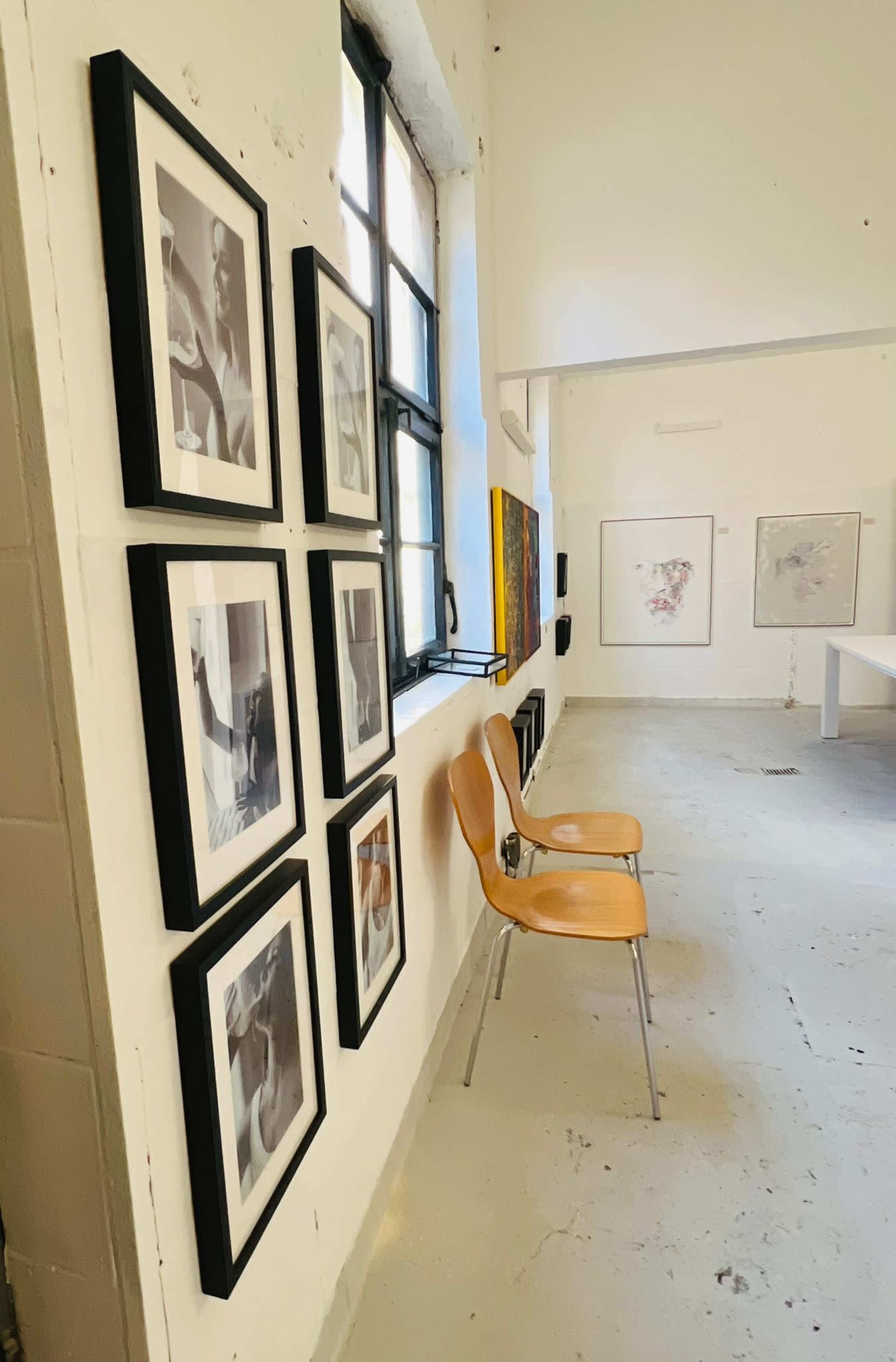 The image shows a gallery interior with a row of framed photographs on the wall and two wooden chairs positioned nearby.