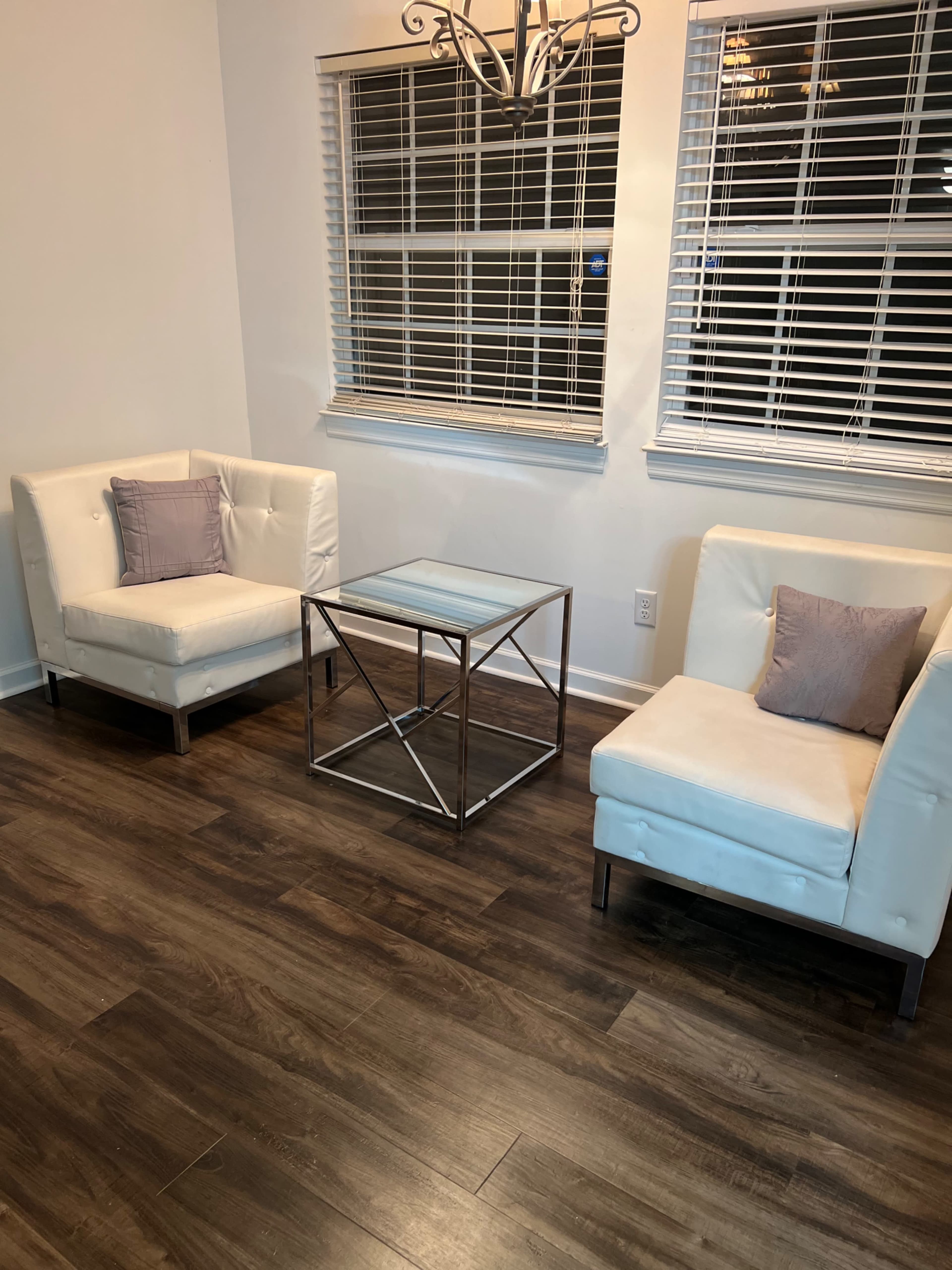 A modern seating area with two white armchairs and a metal coffee table positioned between them, under a chandelier and facing a window with blinds.