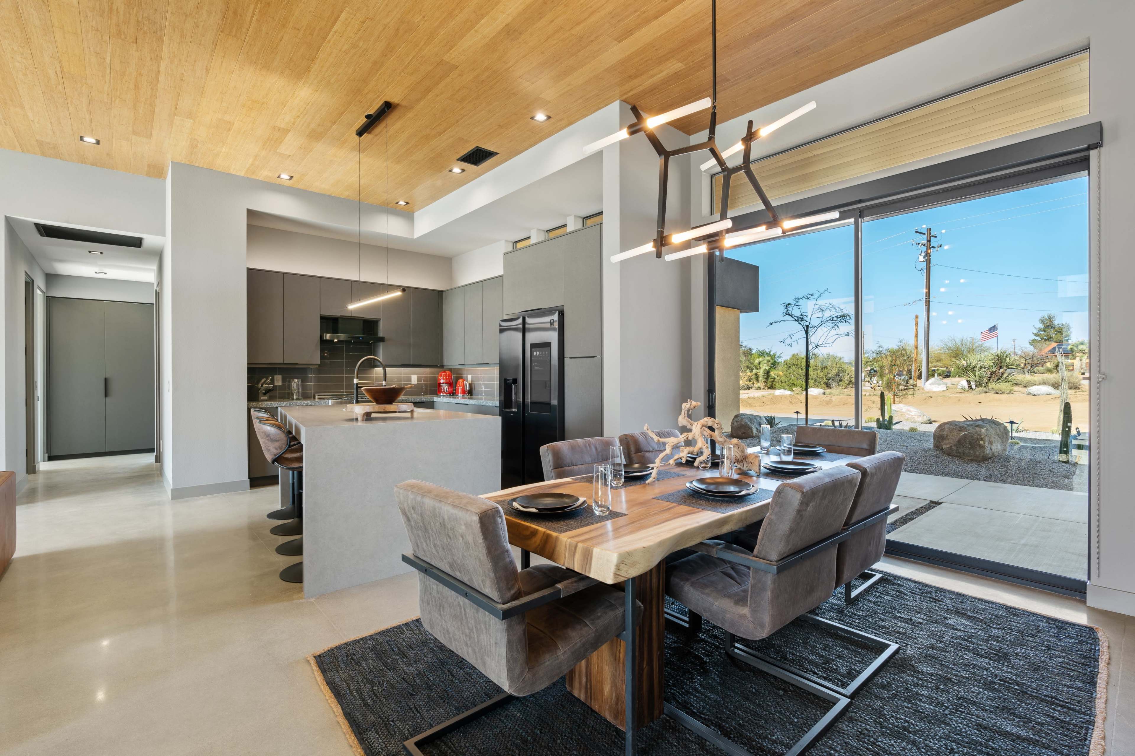 The image shows a modern kitchen and dining area with a wooden ceiling, sleek cabinetry, and a large glass door opening to an outdoor space.