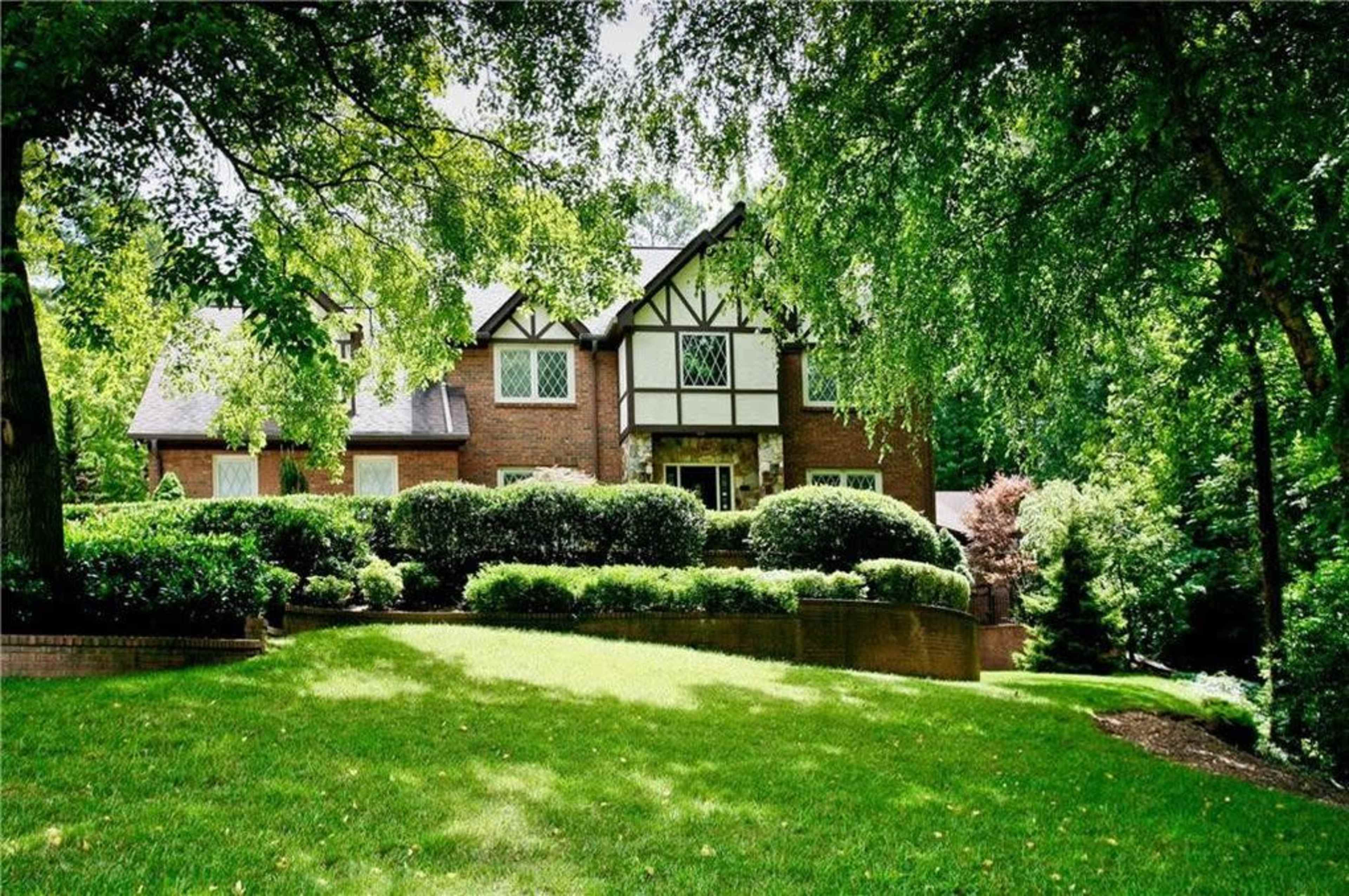 A large brick house with Tudor-style architecture is set on a lush green lawn surrounded by dense trees and neatly trimmed shrubs.