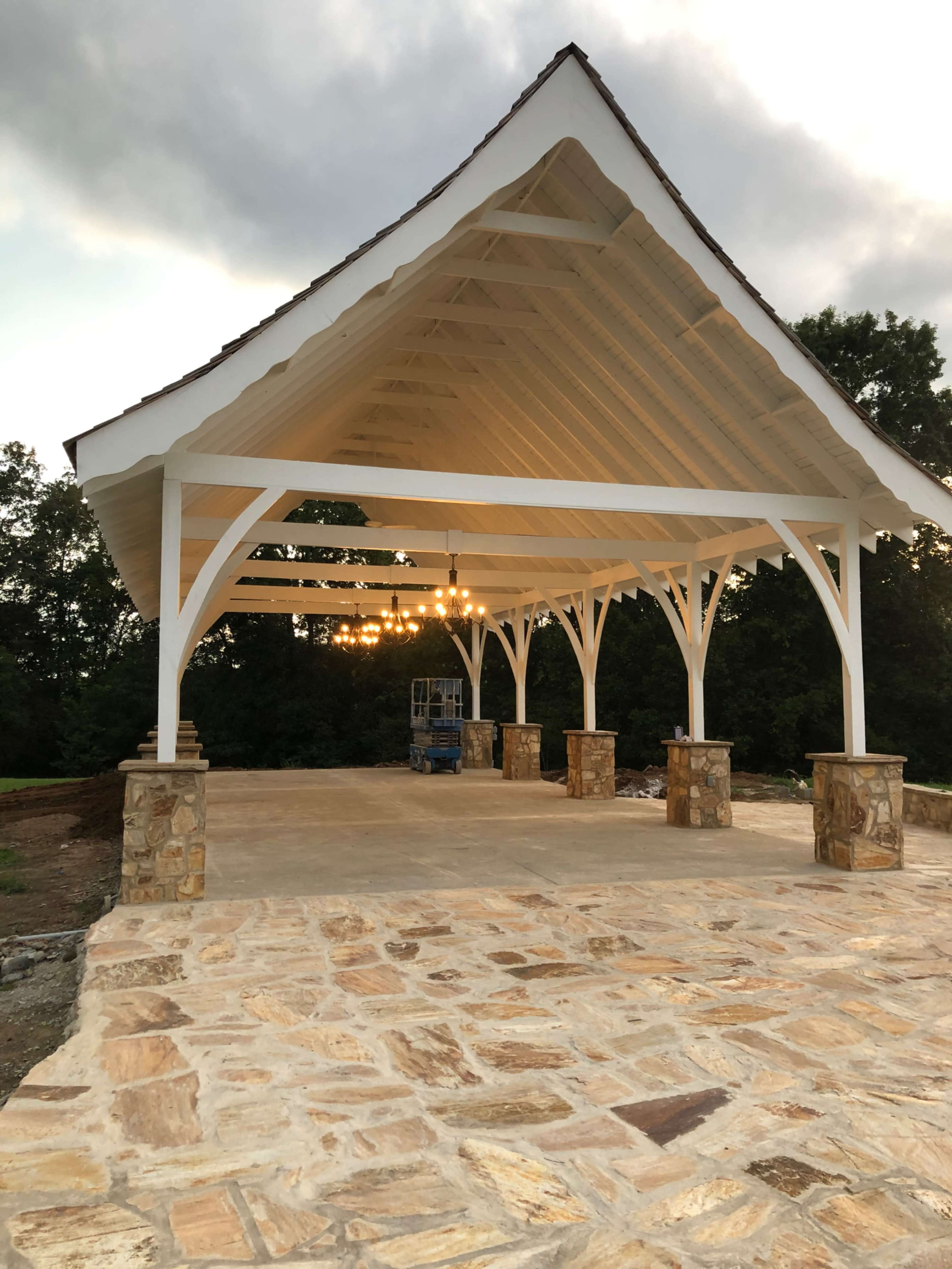 A large, open pavilion with a wooden roof supported by white beams and stone columns, set against a wooded backdrop.