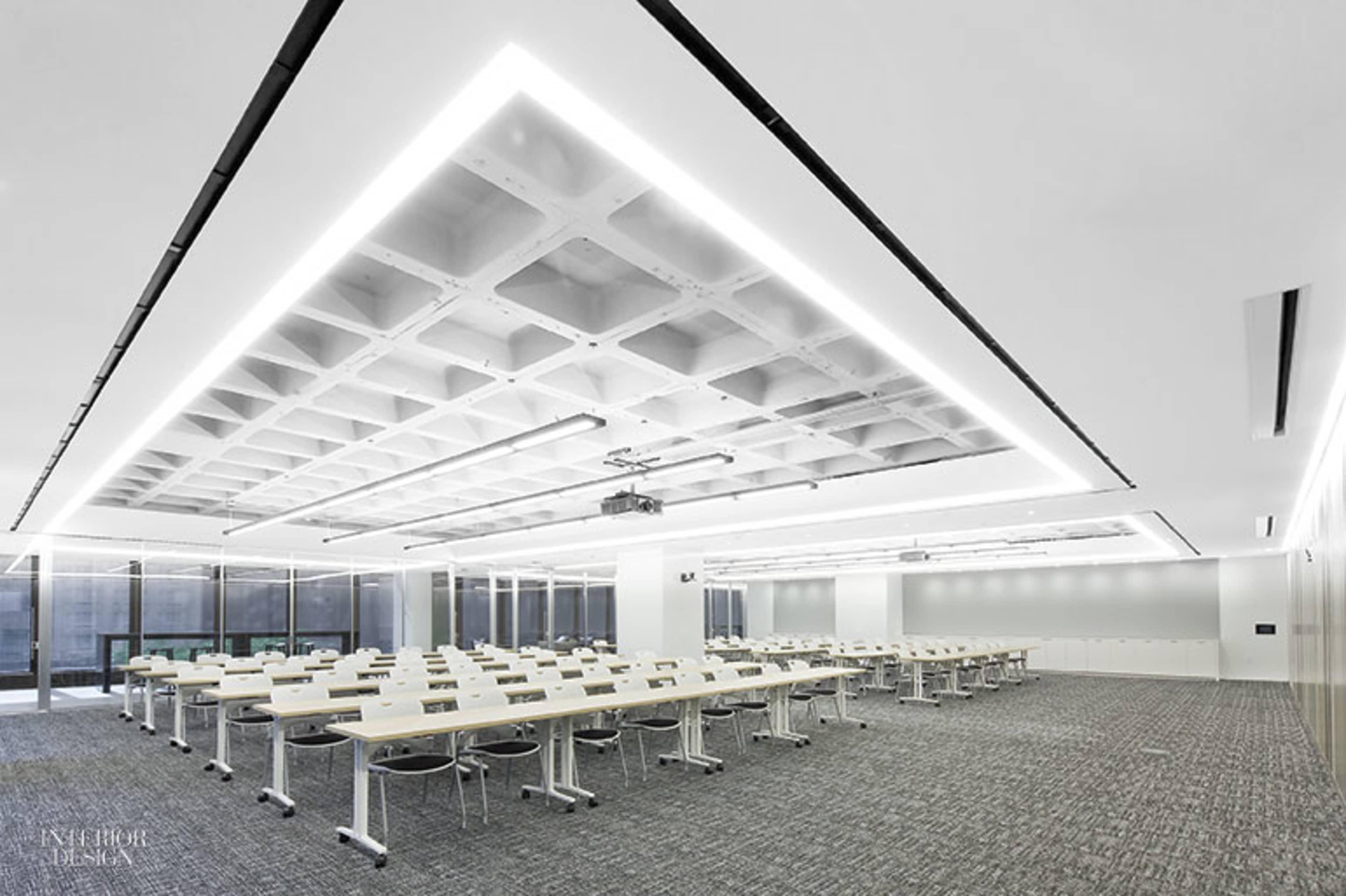 A modern conference room features rows of white tables and chairs under a coffered ceiling with linear lighting.