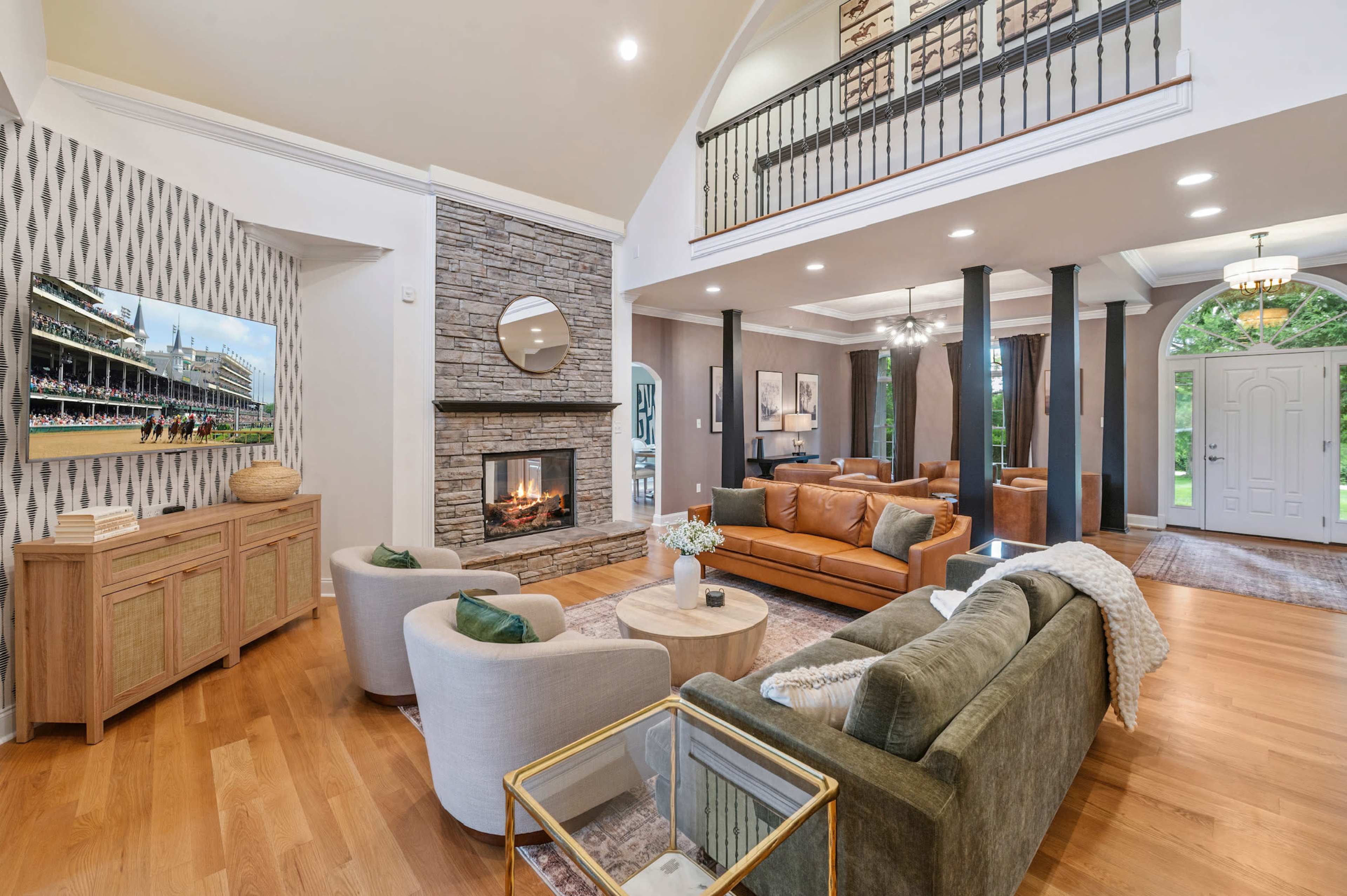 The image shows a spacious living room with a stone fireplace, a large television mounted on the wall, and a mix of seating options including a leather sofa and armchairs, all set under high ceilings.