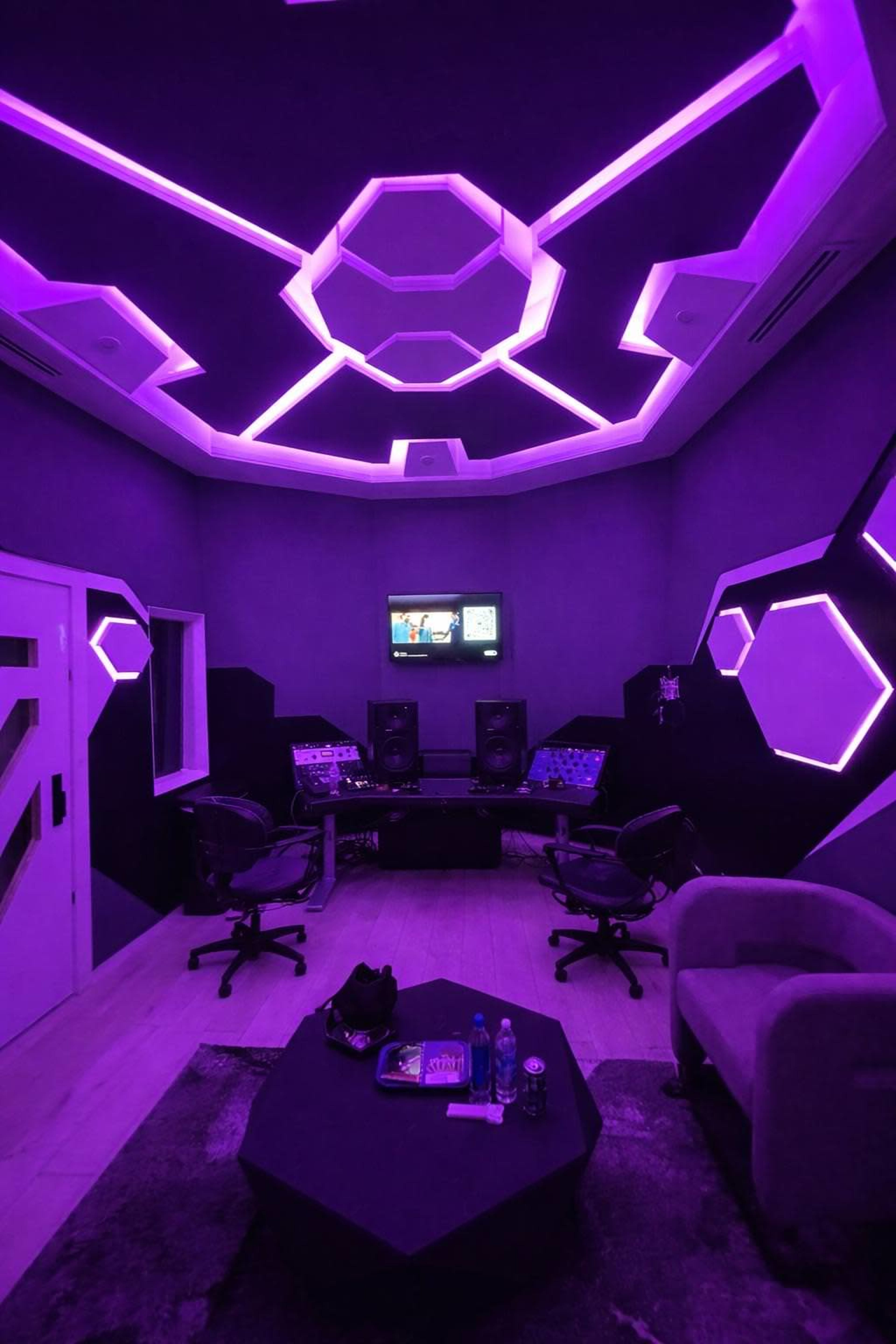 The image shows a modern recording studio with purple LED lights on the ceiling and walls, featuring two chairs, a table, and a monitor on the wall.