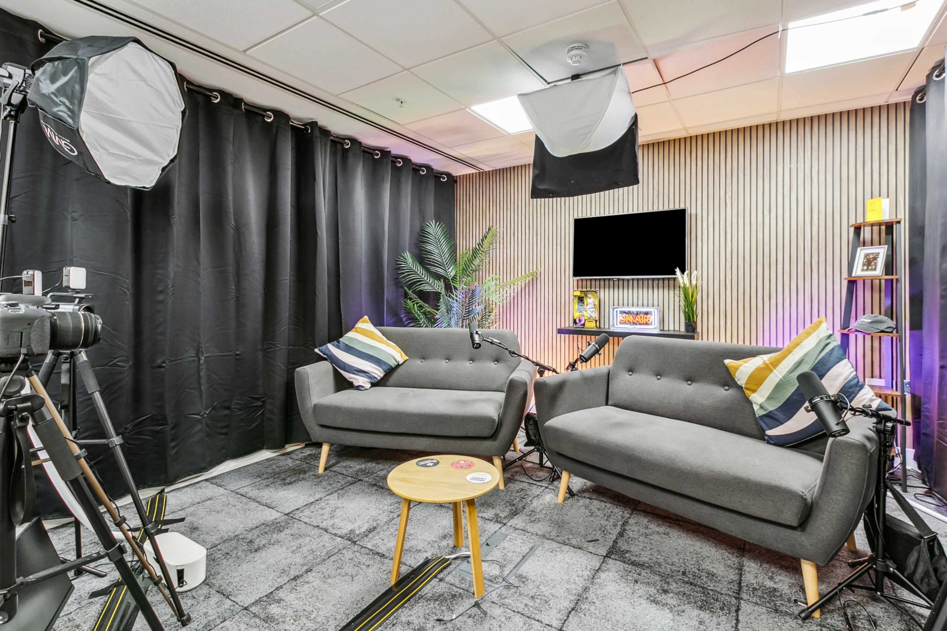 A studio space with two gray sofas, a wooden coffee table, and photography equipment in front of a backdrop of black curtains and a wooden-paneled wall.