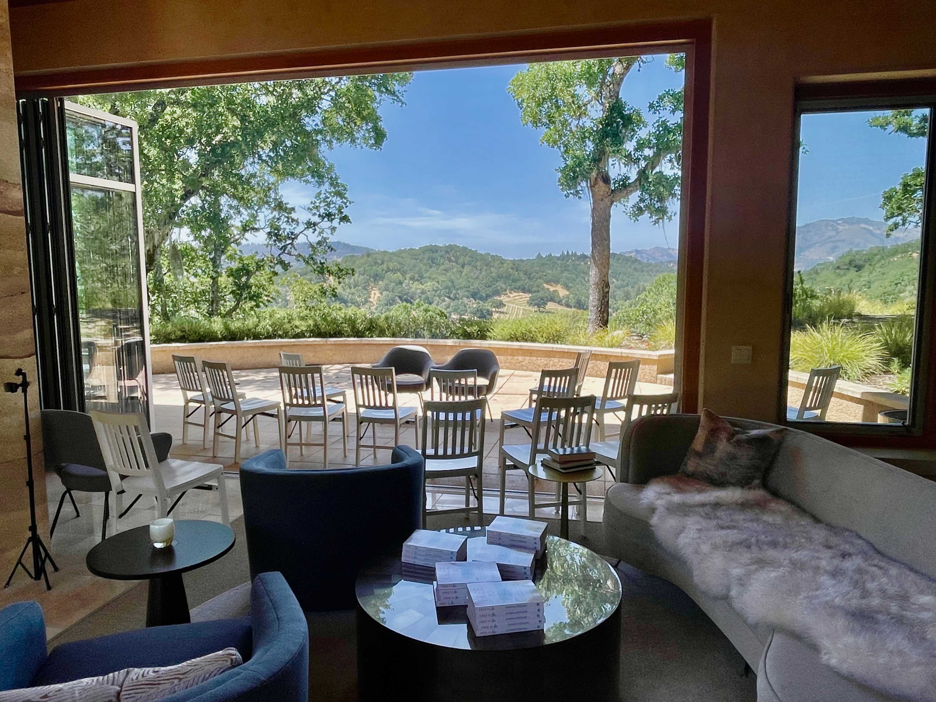 A spacious room with a modern seating area opens to a panoramic view of hills and trees through large glass doors.