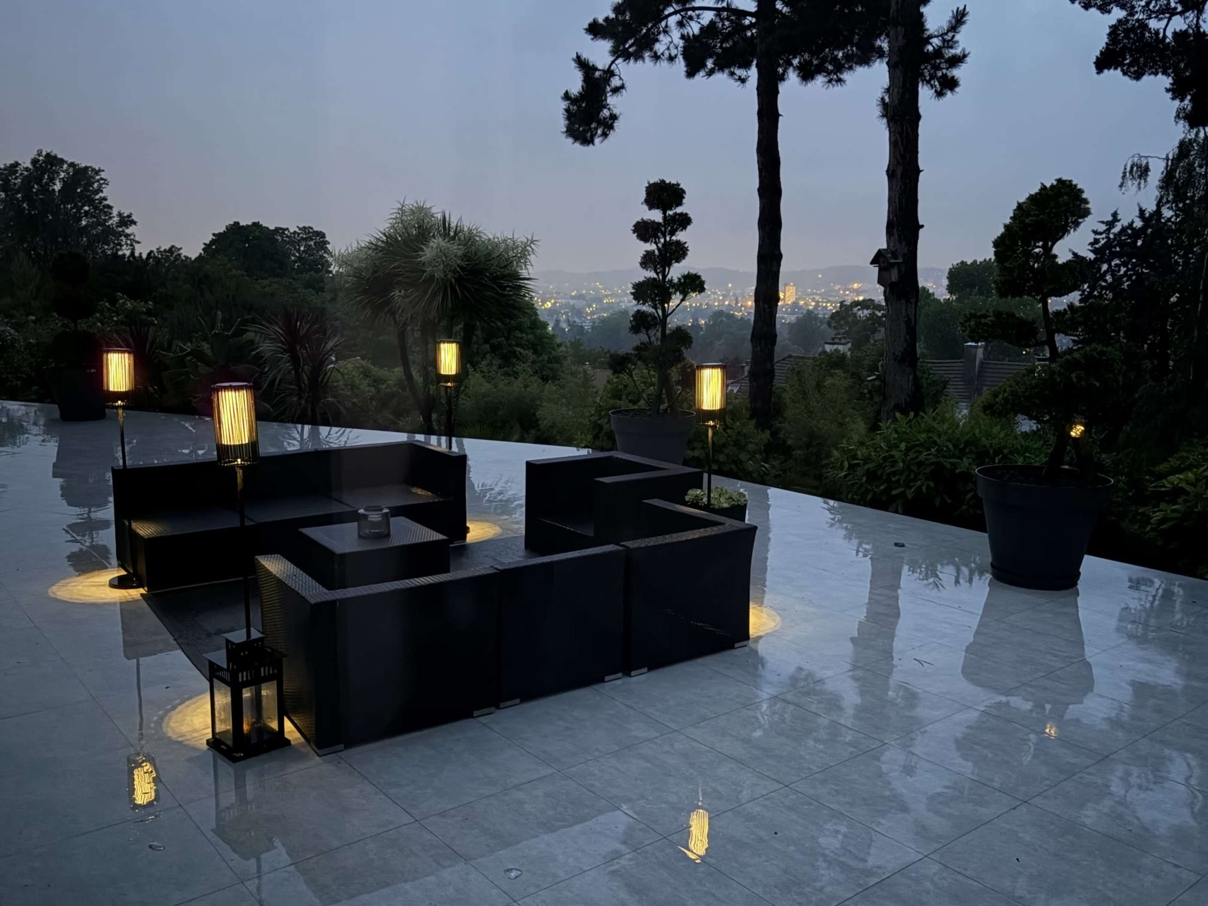 The image shows a modern outdoor lounge area with black seating, illuminated lanterns, and a view of distant lights through a faint twilight mist.