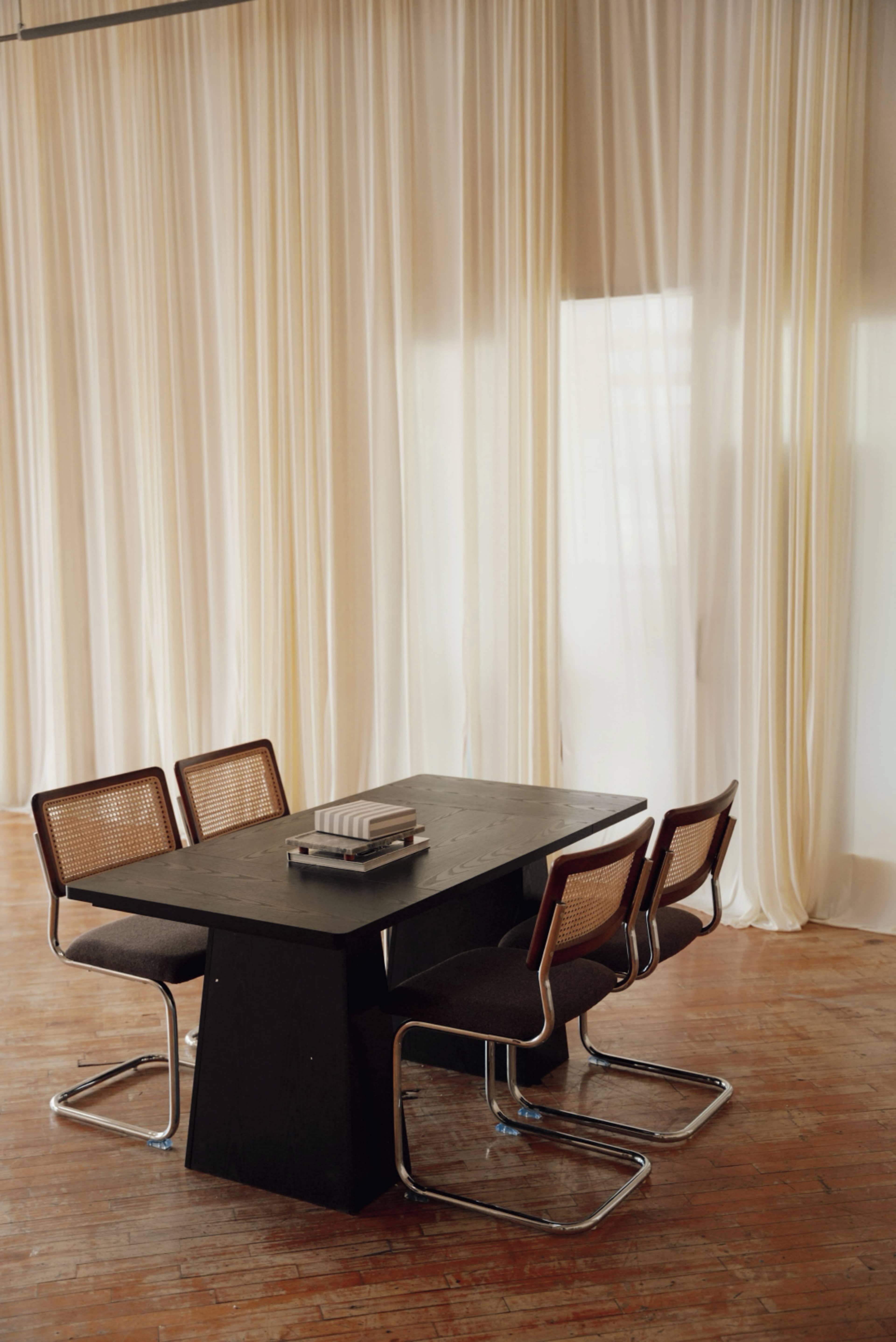 A modern dining table is accompanied by four chairs, set against a backdrop of sheer, light-colored curtains.