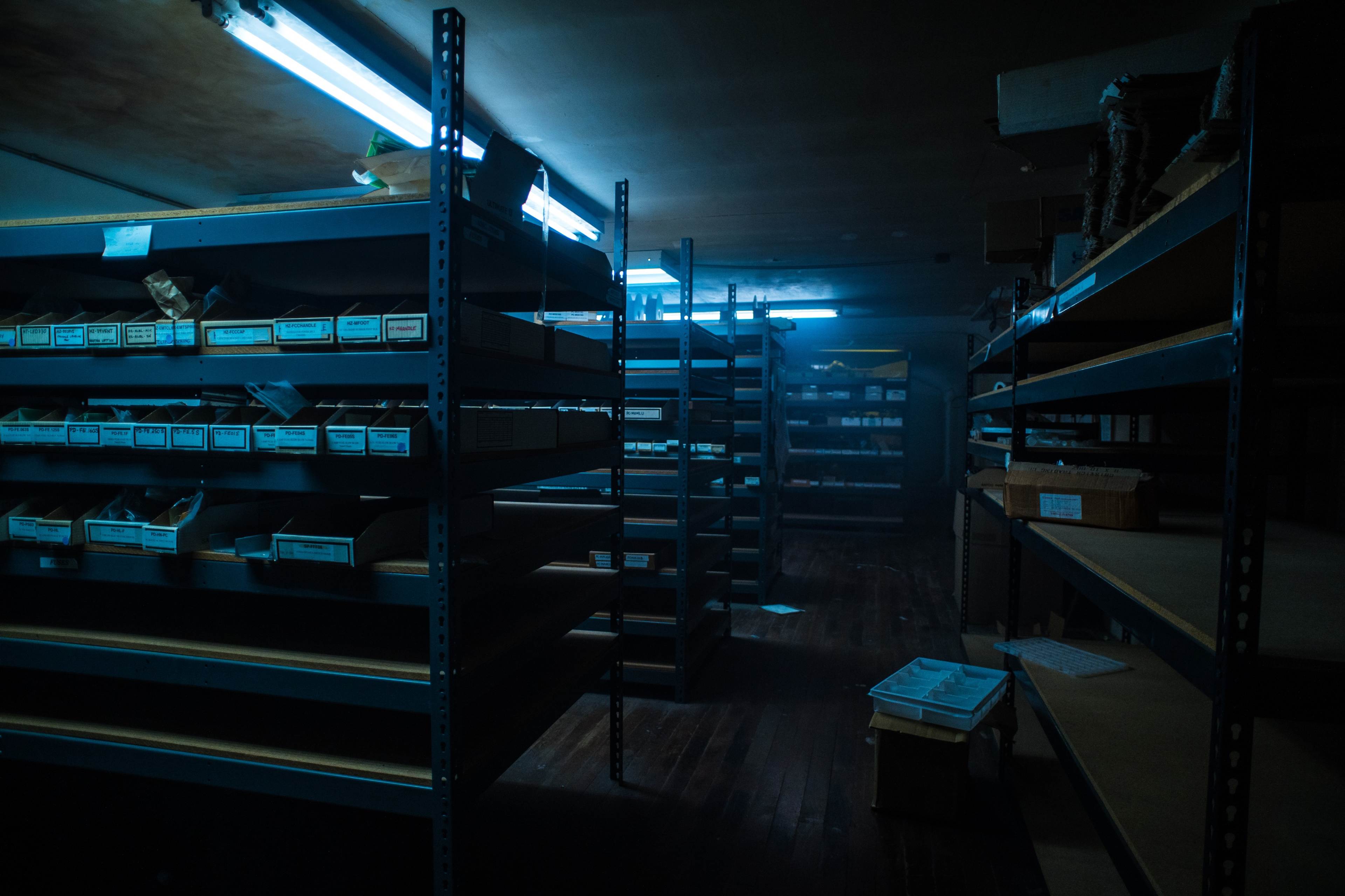A dimly lit storage room features metal shelves filled with labeled boxes and illuminated by fluorescent lights.