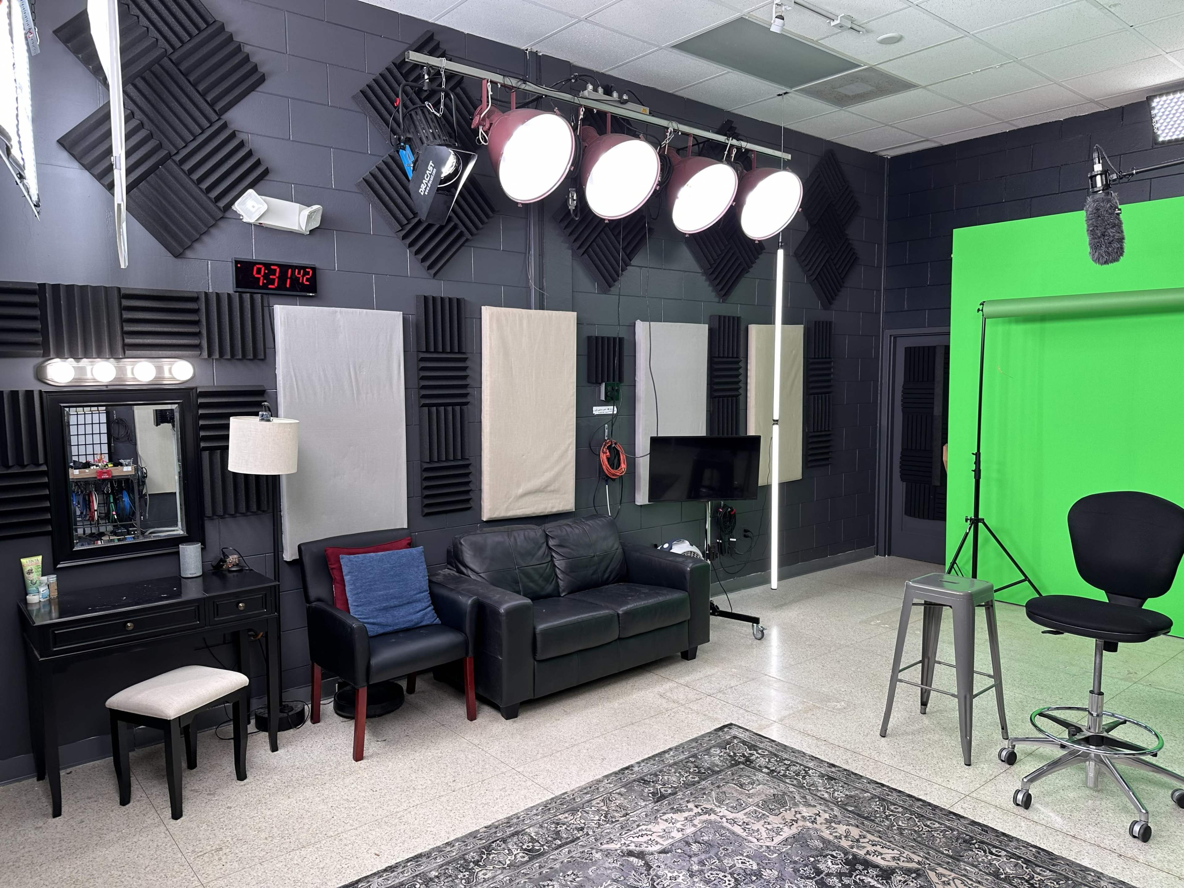 The image shows a studio setup featuring a green screen, various lighting fixtures, soundproofing panels, a black couch, and styling furniture.