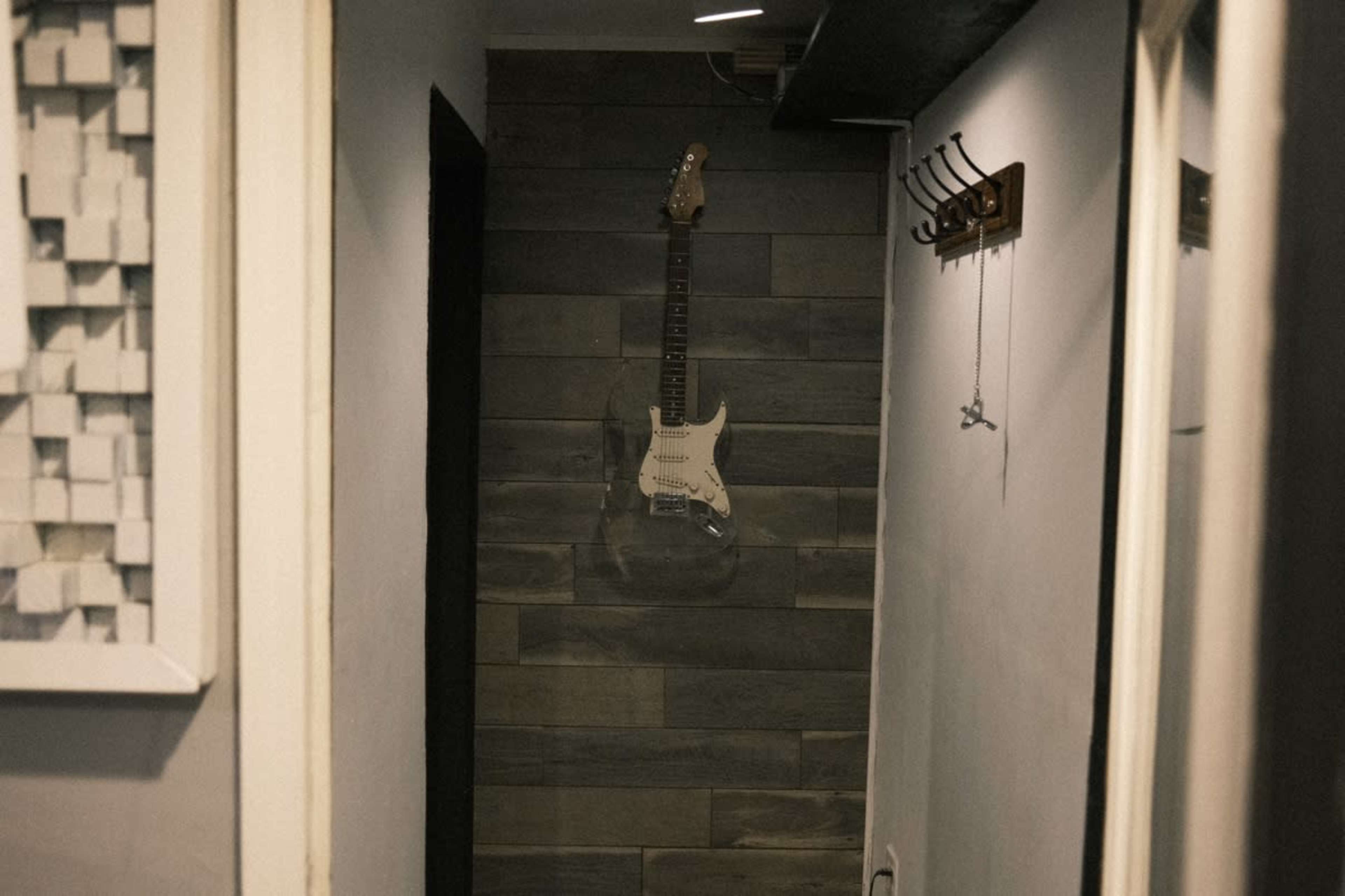 A guitar hangs on a wall in a narrow hallway, flanked by a coat rack and a mirror.