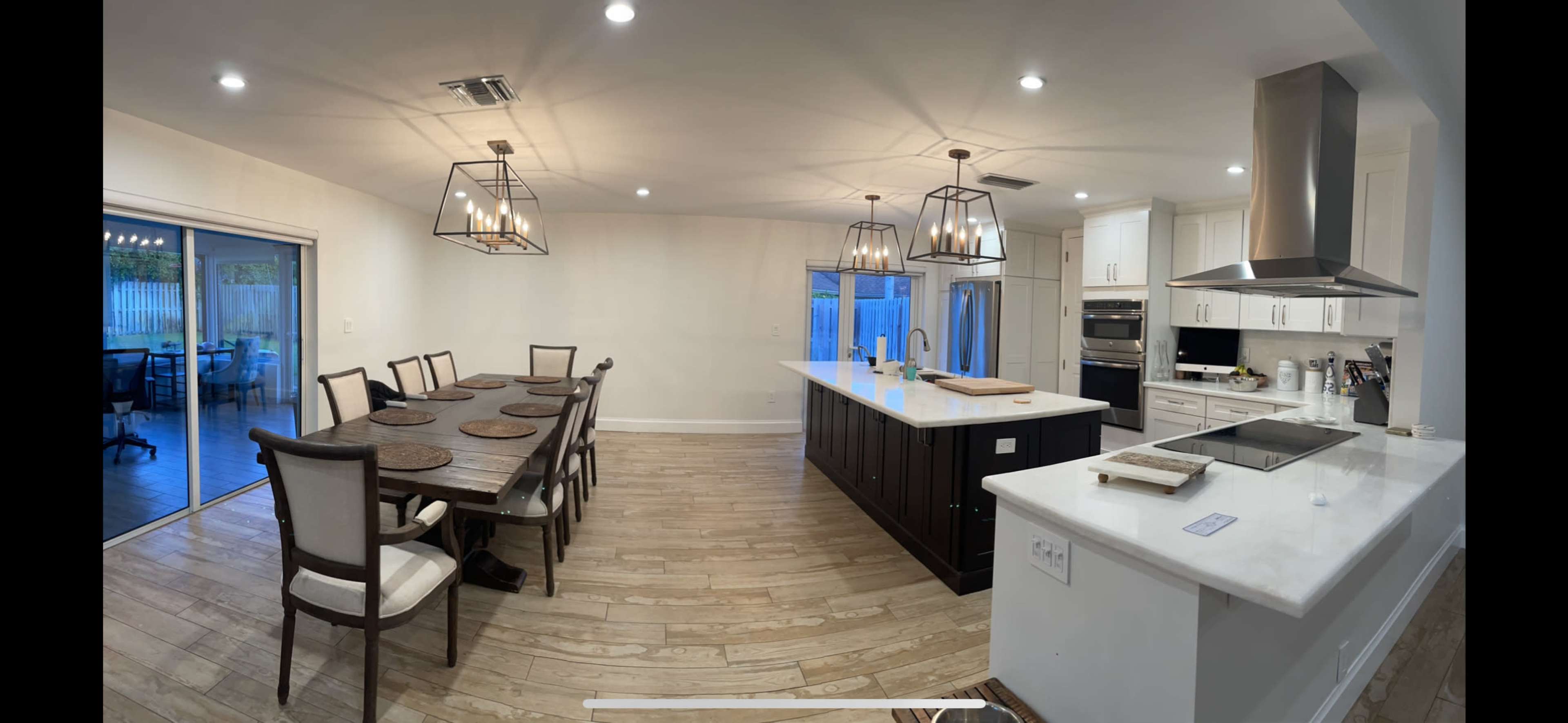 The image shows a modern kitchen and dining area featuring a long dining table with chairs, an island with seating, and large windows facing an outdoor space.