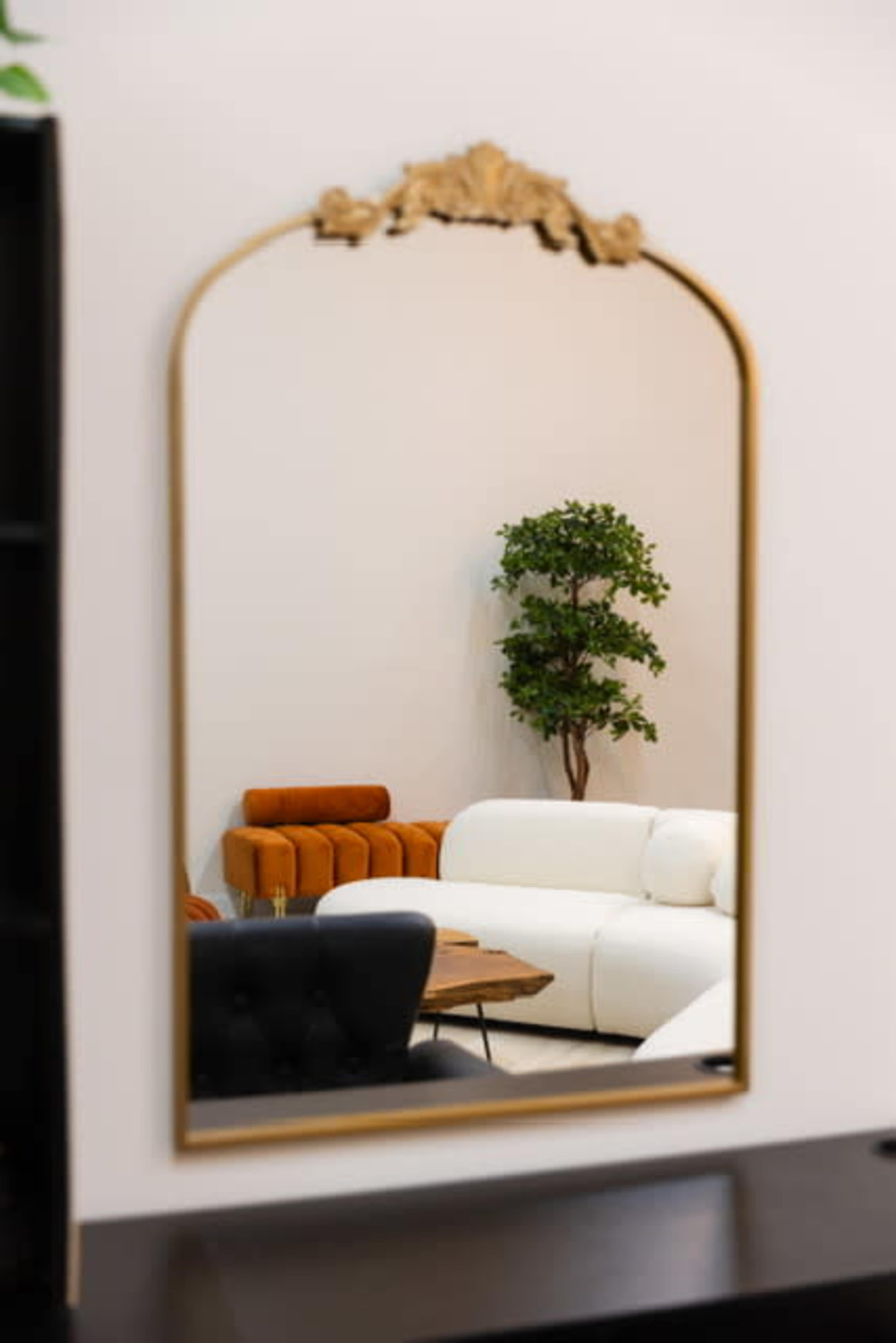 The image shows a decorative mirror reflecting a living room with a white sofa, an orange armchair, and a small green plant.