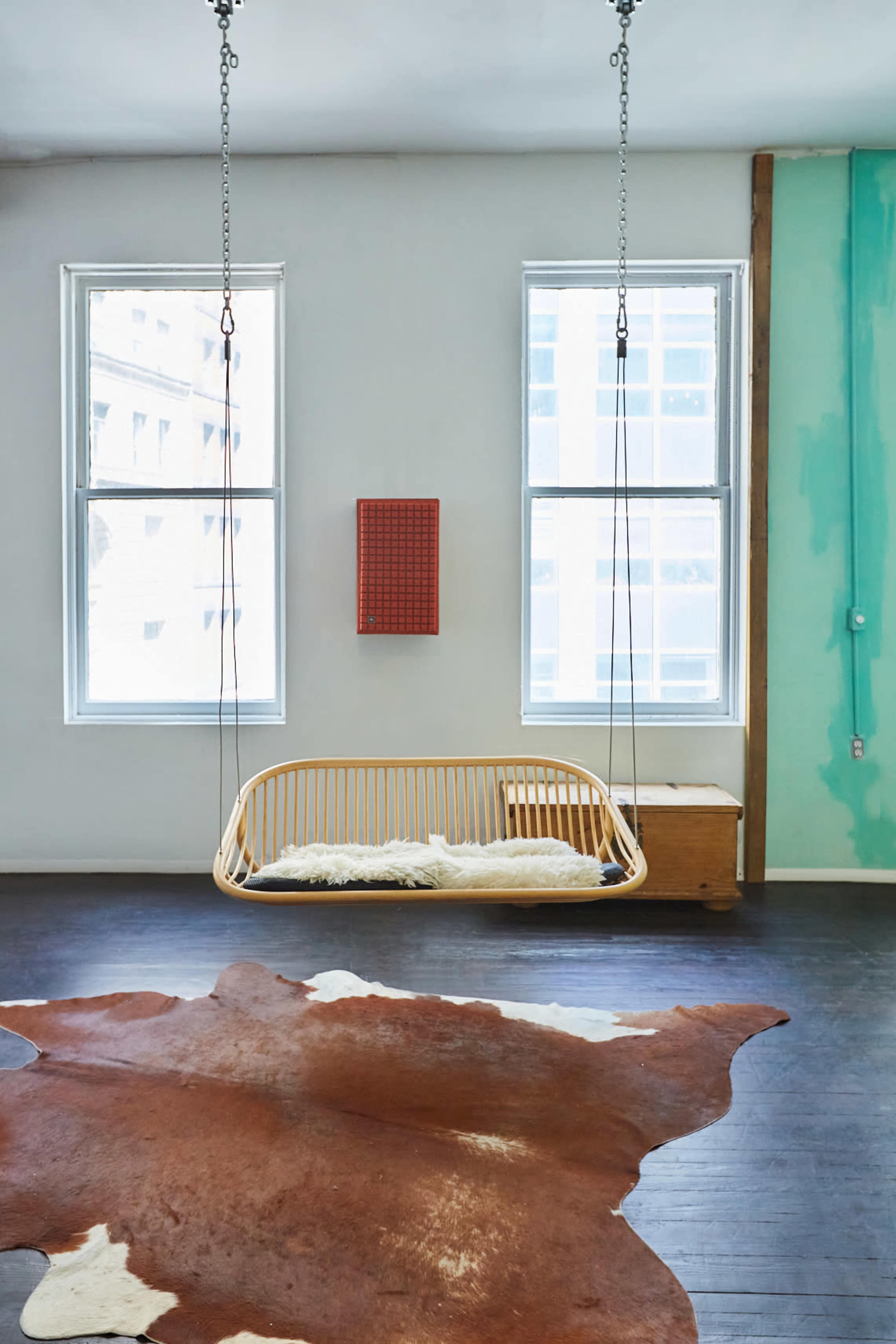 A wooden swing chair hangs from the ceiling in a room with two large windows and a cowhide rug on the floor.