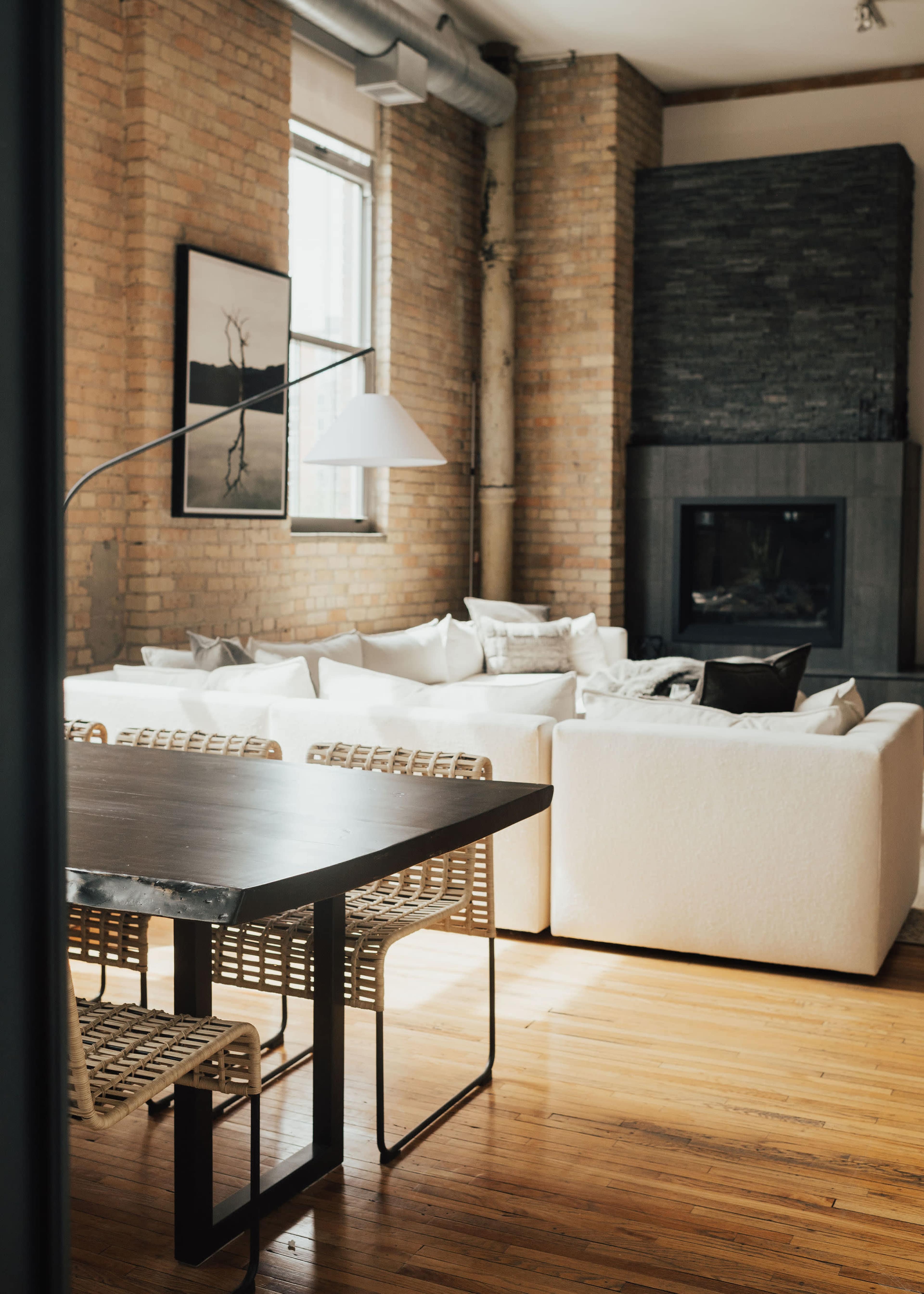A contemporary living room features a white sectional sofa, a wooden dining table with woven chairs, large windows, and a stone fireplace.