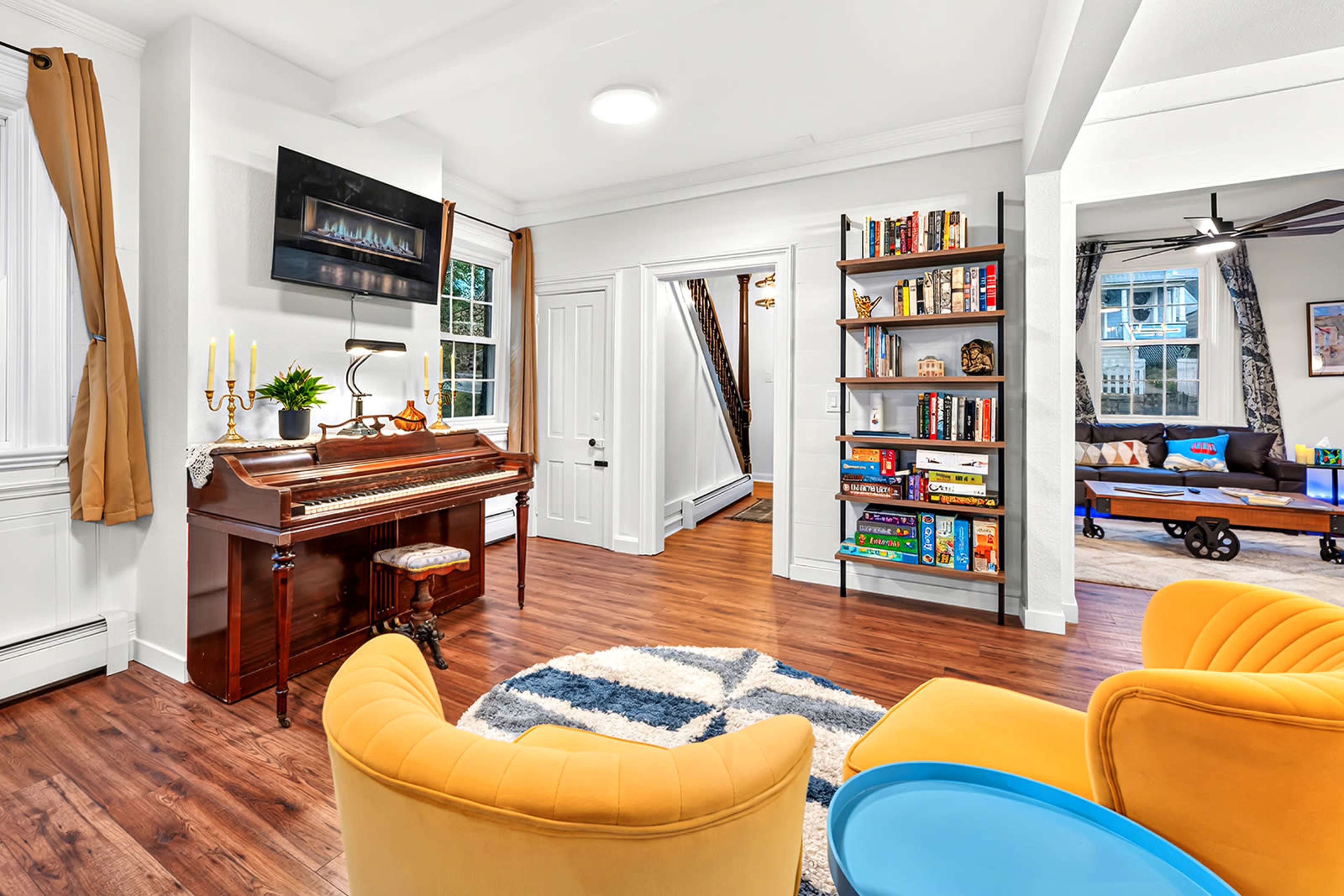 The image shows a cozy living area featuring a piano, a bookshelf filled with books and games, and two yellow armchairs arranged on a patterned rug.