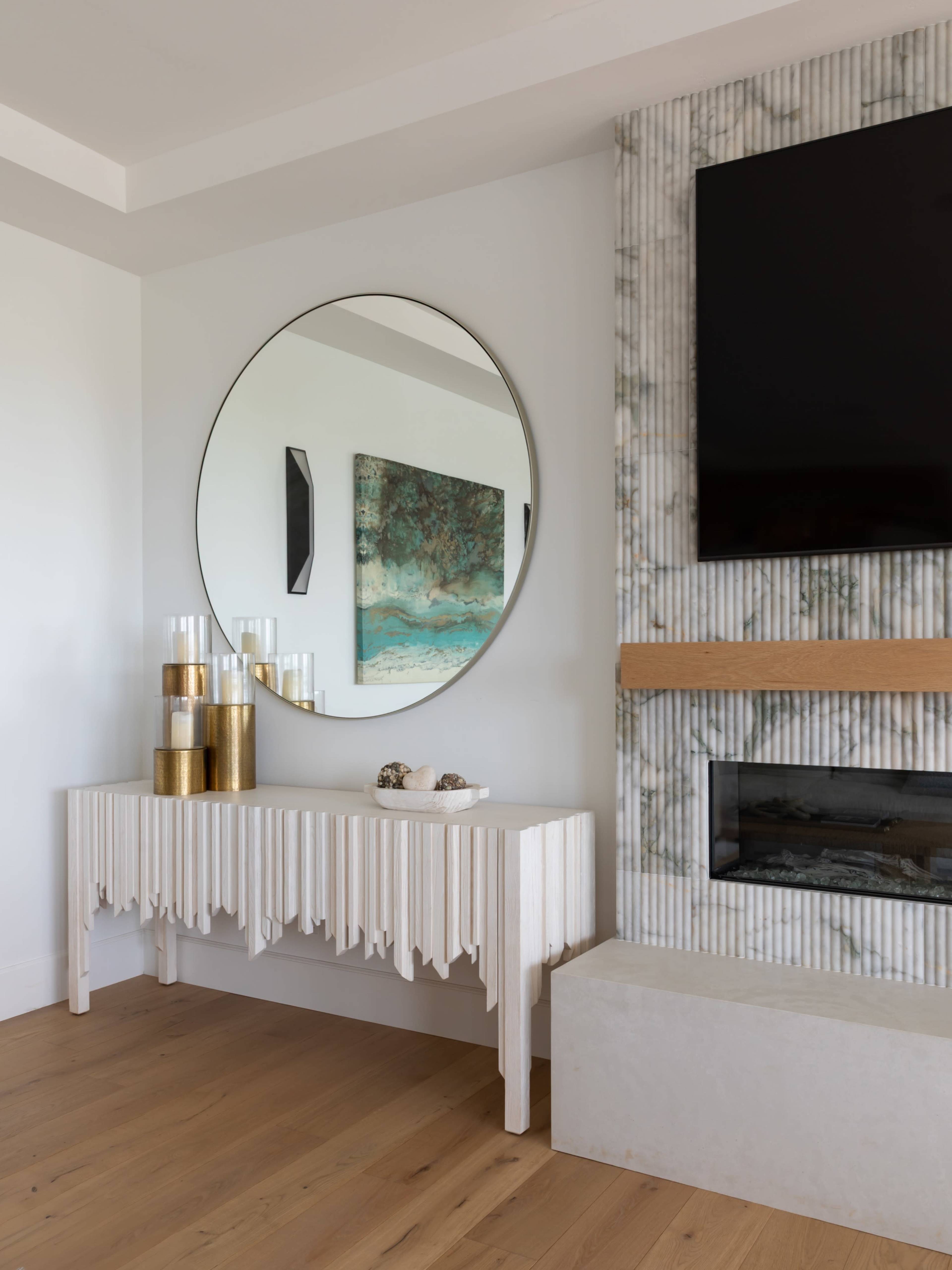 A wooden console table with a textured front is placed beneath a large round mirror and next to a wall-mounted television, with a decorative piece and candles on the table.