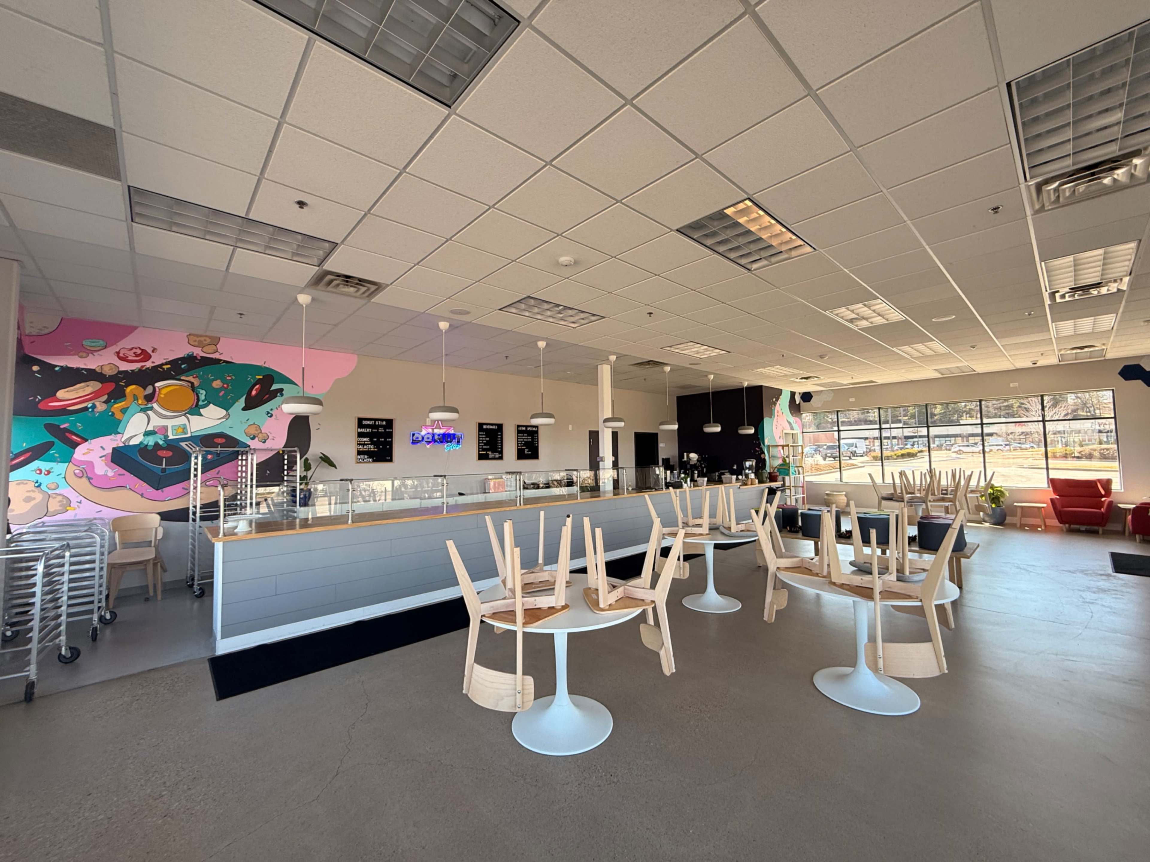 The interior of a modern café features empty chairs stacked on tables, a large counter with a display case, and colorful wall art on one side.