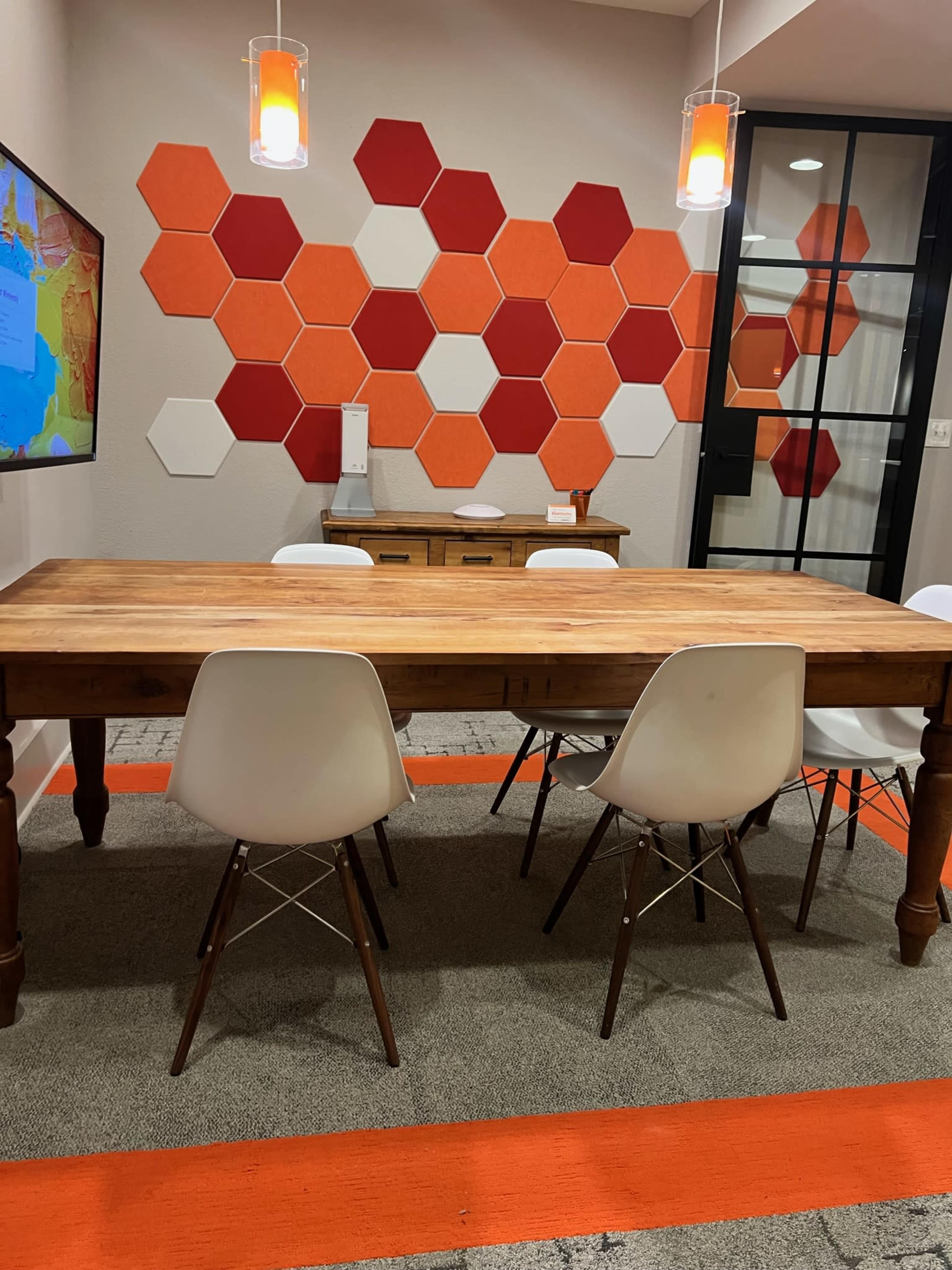 A wooden table surrounded by white chairs is set against a wall adorned with red and orange hexagonal tiles.