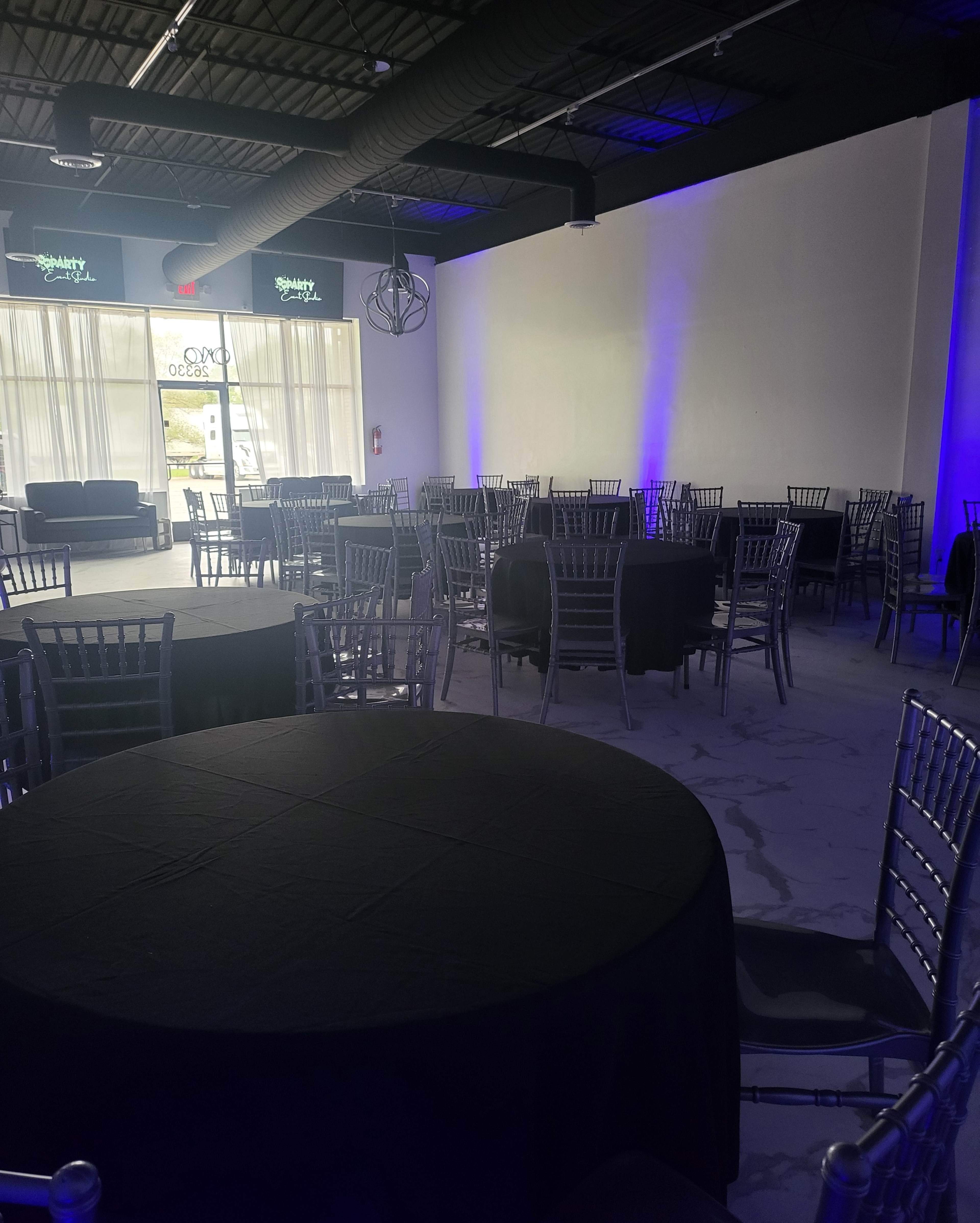 A spacious event hall with several round tables covered in black cloth and silver chairs arranged throughout the room, illuminated by blue lighting.