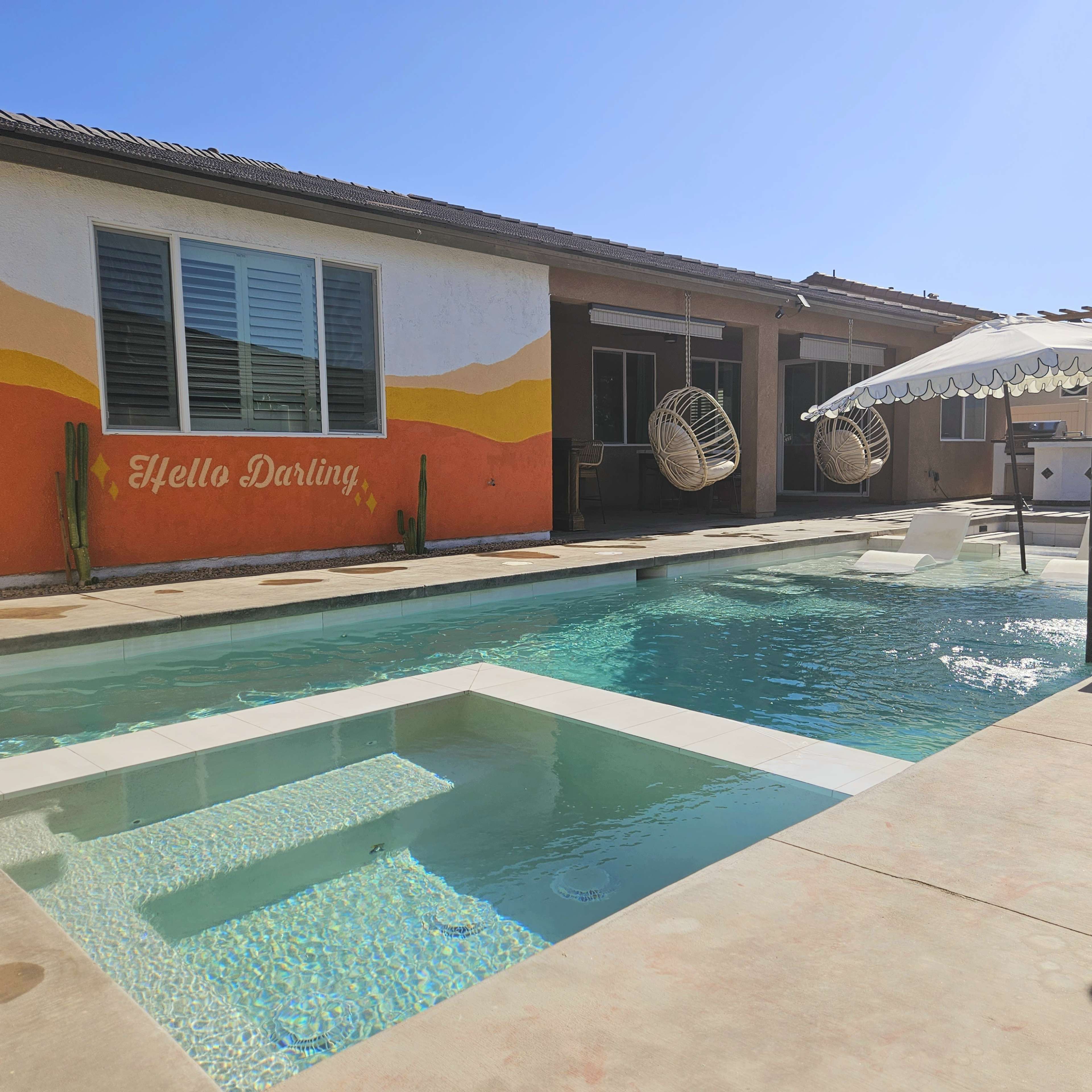 A swimming pool with a spa area, surrounded by a patio and a wall with the words "Hello Darling" painted on it.